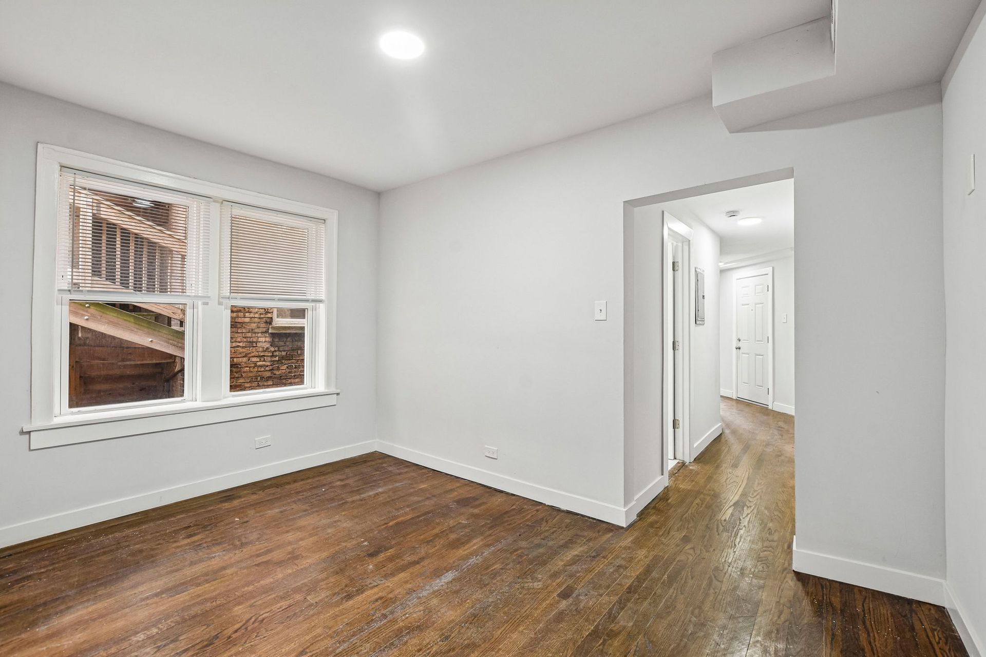 Empty white room with hardwood floors, recessed lighting, and a window facing a hallway.
