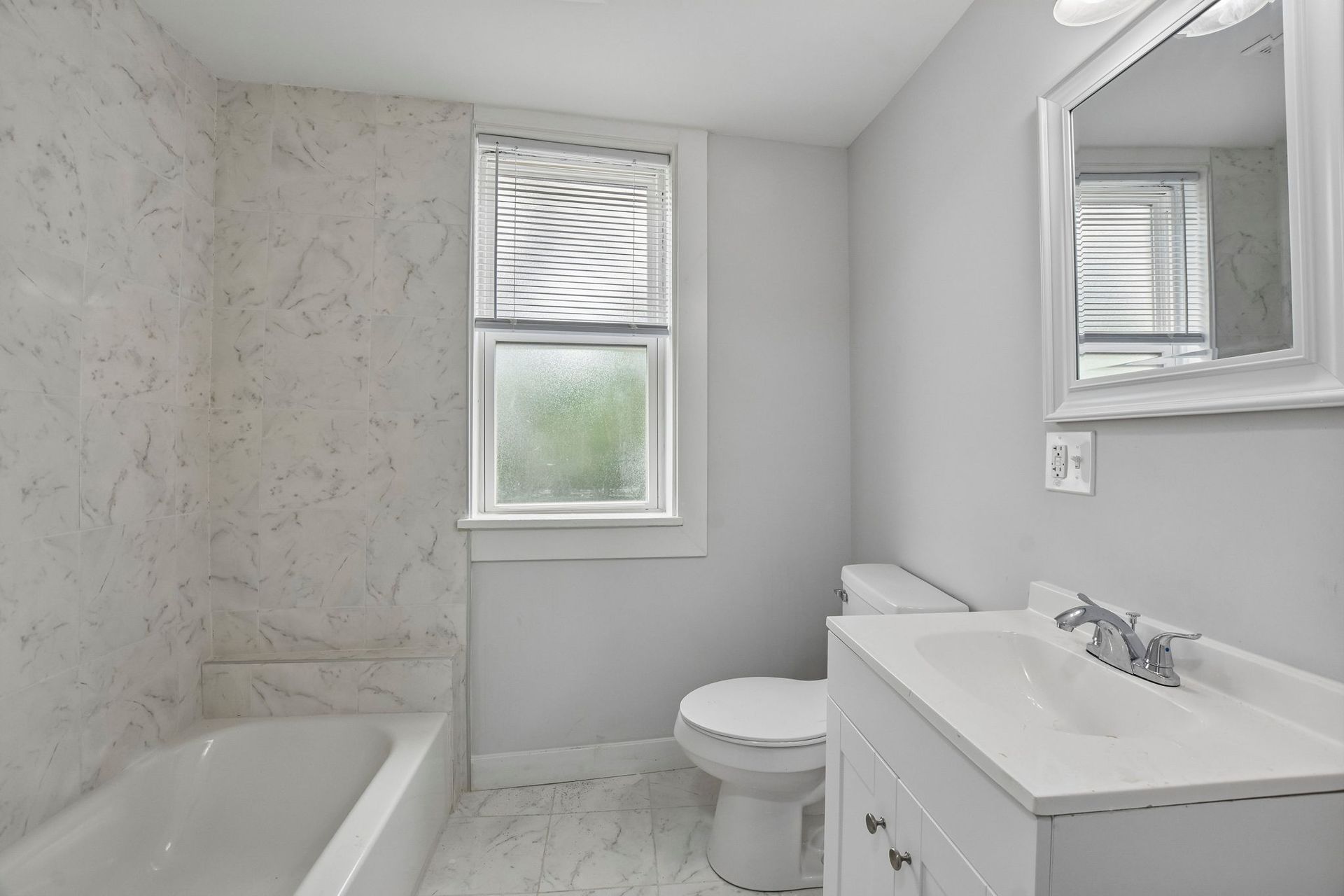 Bright white bathroom with bathtub, toilet, vanity sink, and small window.