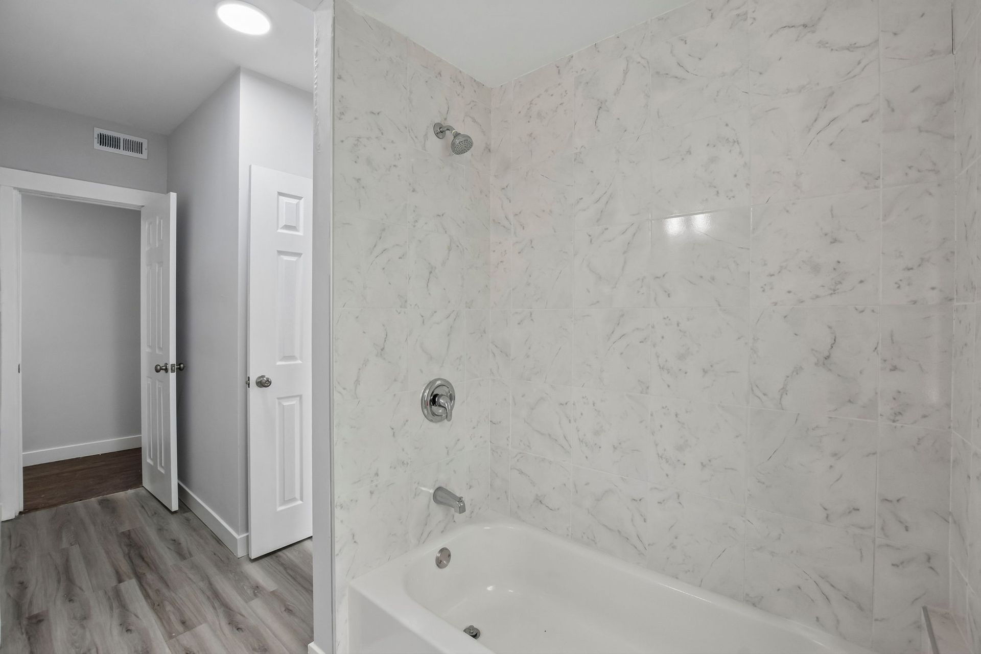 Bright white bathroom with marble-look tub surround, gray wood floor, and hallway visible through open doors