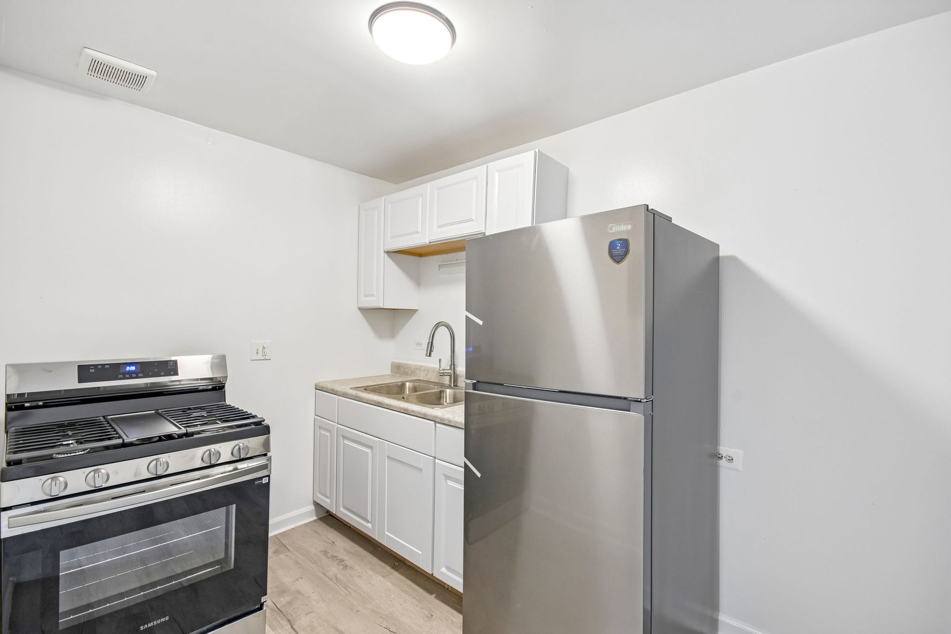 Small white kitchen with stove, sink, and stainless steel refrigerator under bright ceiling light