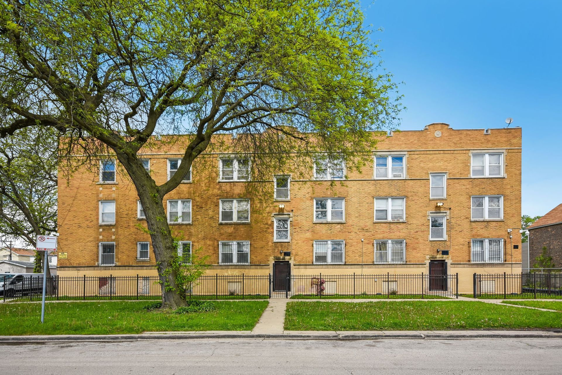 Brick apartment building behind a large tree and green lawn under a cloudy sky