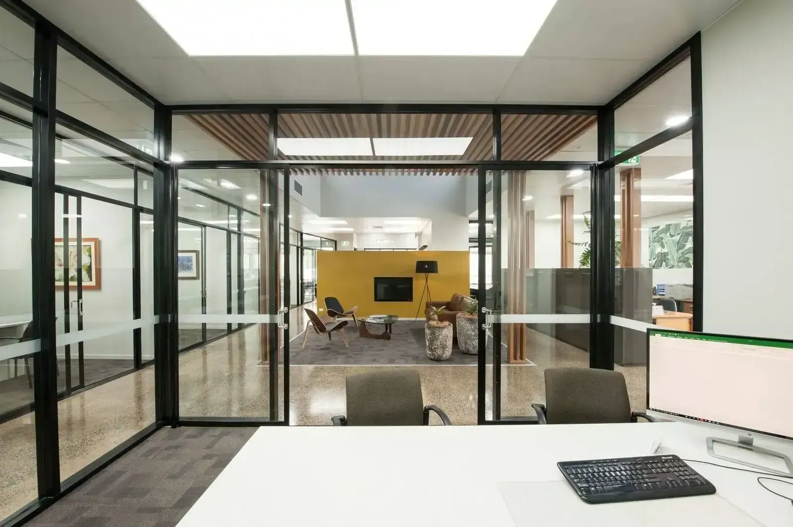 A modern office interior viewed from a desk, looking through glass walls toward a lounge area with yellow feature walls. — Downs Window Renewals in Harristown, QLD