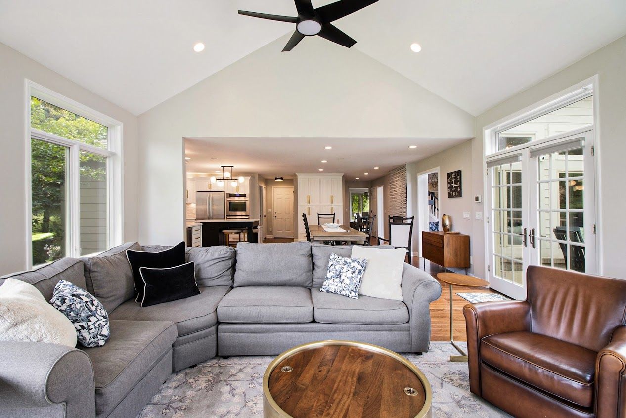 Bright living room with gray sectional, brown leather chair, open kitchen, and large windows