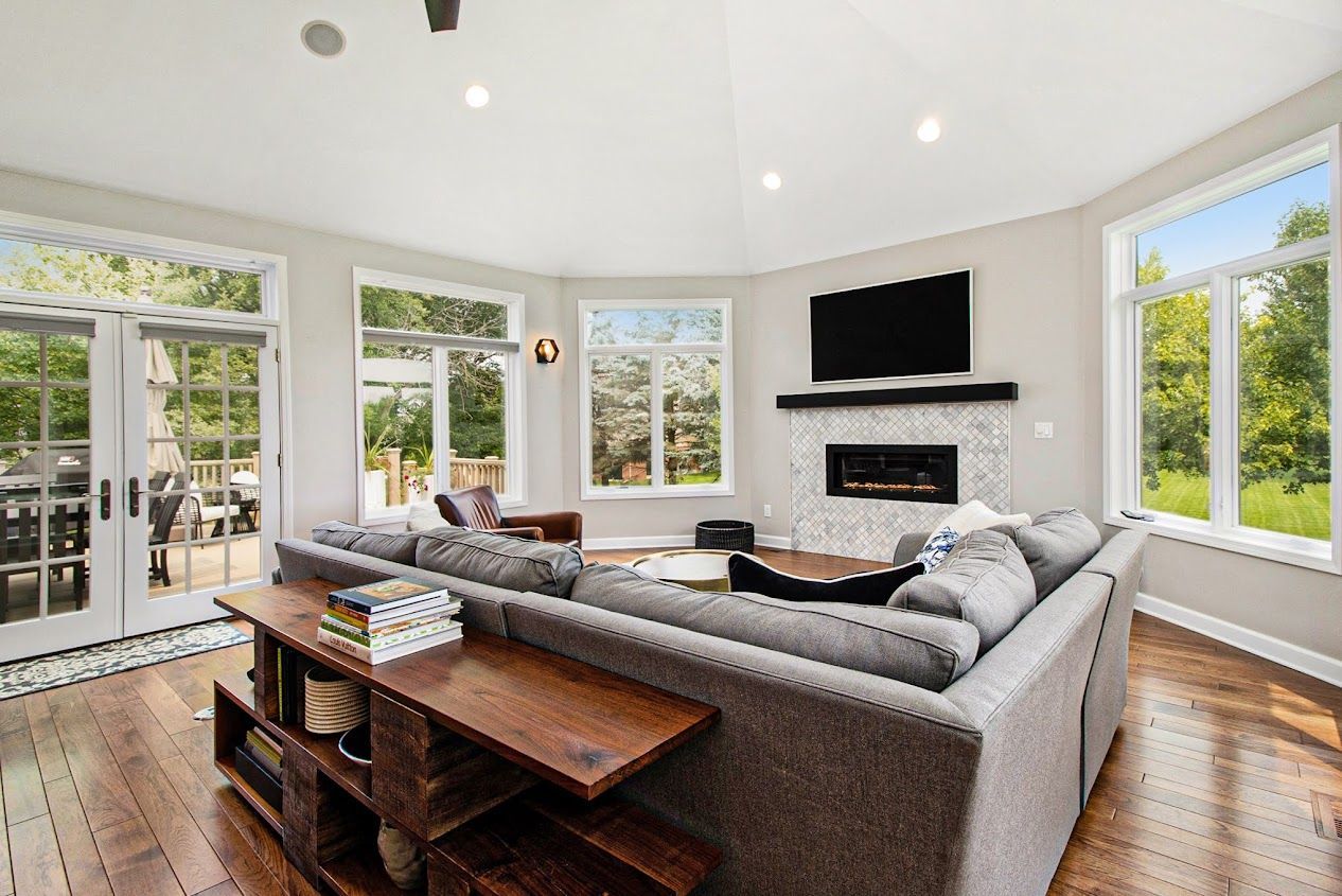 Bright modern living room with gray sectional, wall-mounted TV, fireplace, large windows, and wooden floors
