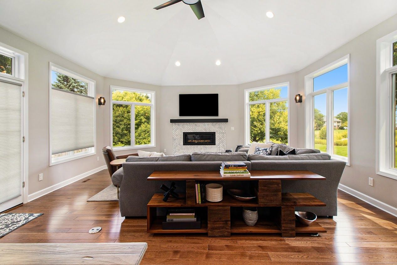 Bright living room with large windows, hardwood floors, gray sofa, and wall-mounted TV above a fireplace.