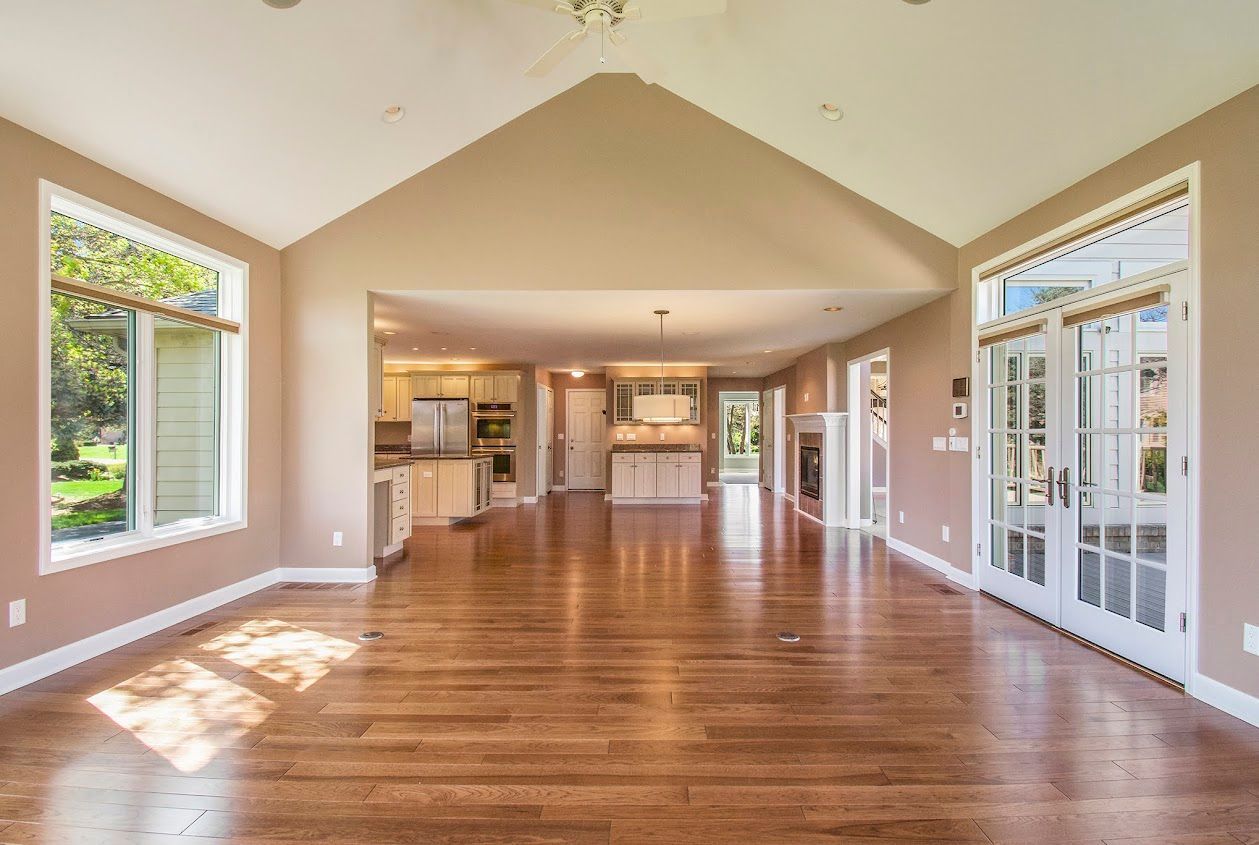 Spacious empty living room with hardwood floors, high ceiling, and large windows letting in sunlight.