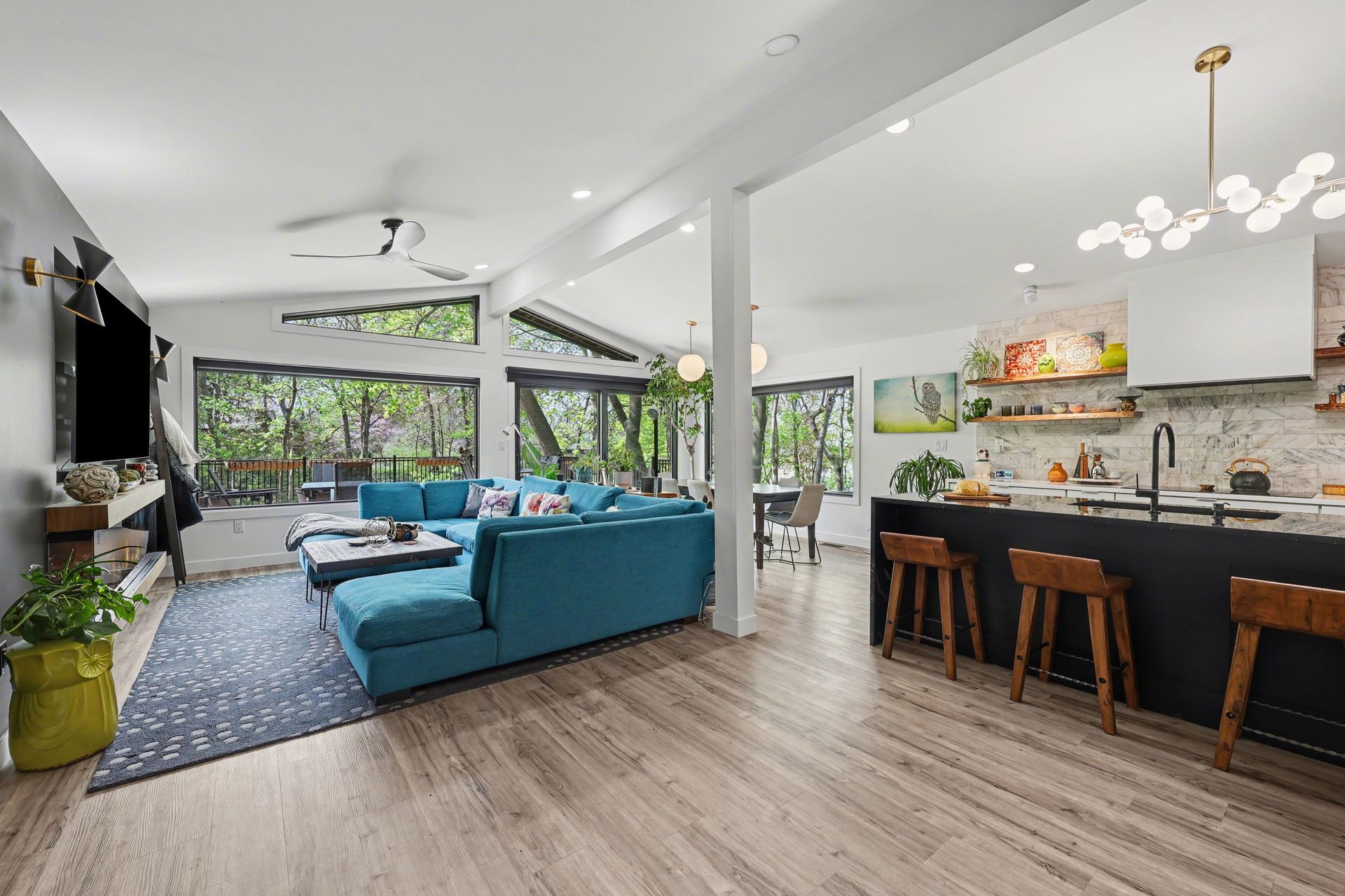 Bright open living room with blue sofa, kitchen island stools, and large windows overlooking greenery