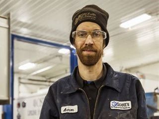 Mechanic in work uniform and safety glasses, wearing a beanie. Standing in a garage, smiling.