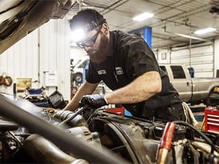 Mechanic working on car engine, wearing headlamp and gloves in a repair shop.