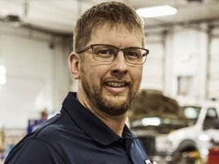 Man with glasses and beard in auto shop setting, smiling at camera.