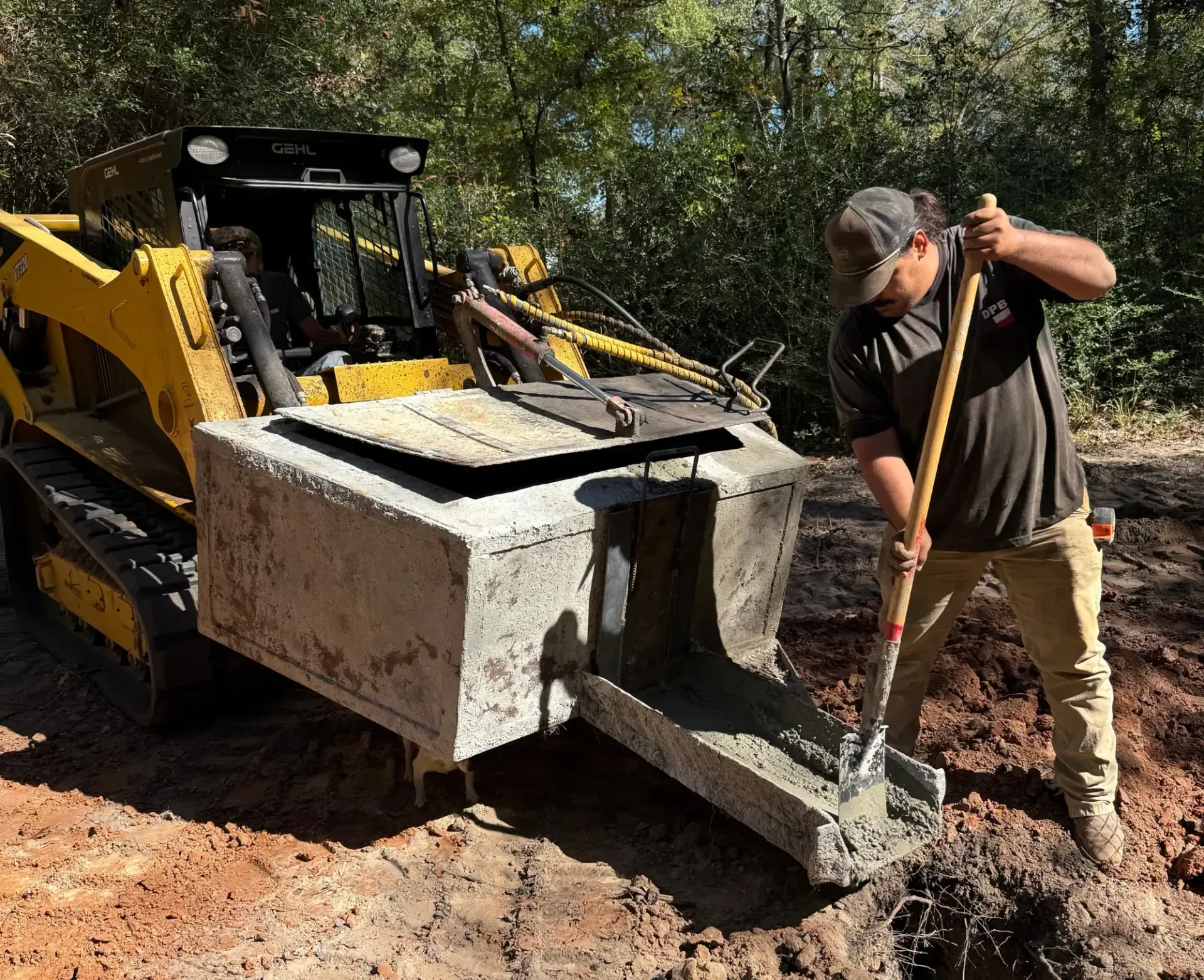 Man using a shovel beside a yellow skid steer with a front bucket in muddy woods