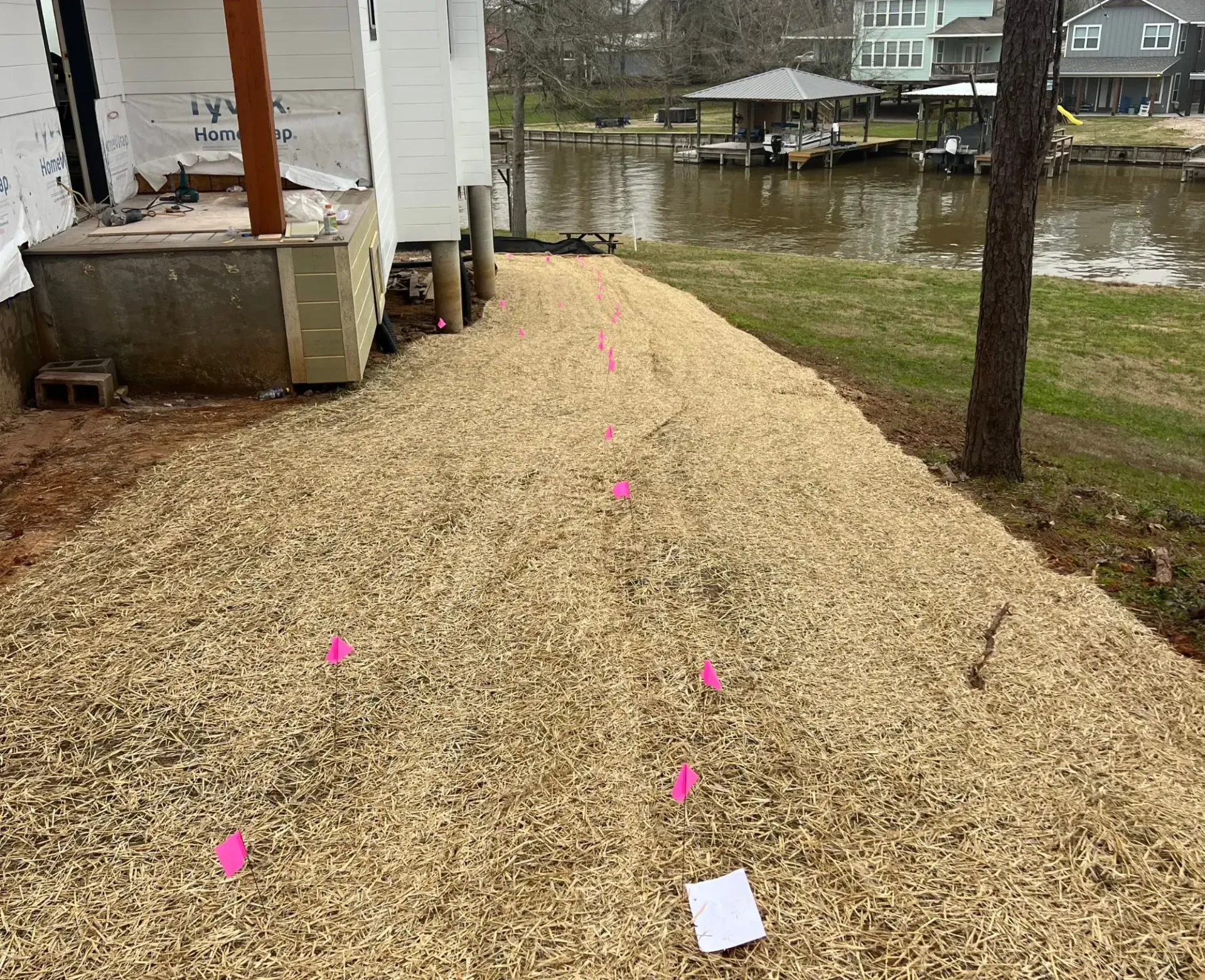 Fresh gravel path beside a house, marked with pink flags, beside a flooded yard and lake.