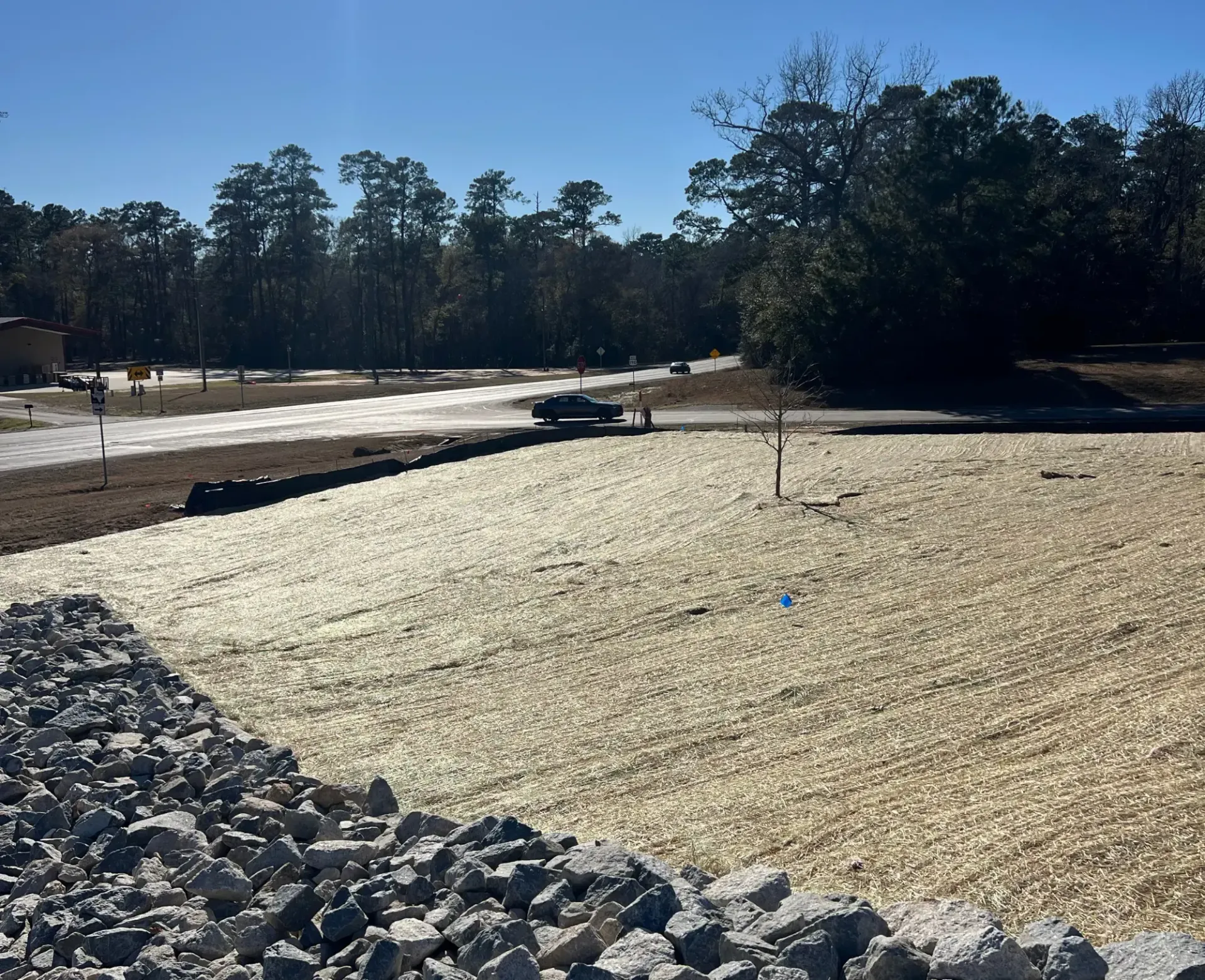 Construction site with a rocky embankment, paved area, and trees under a clear blue sky