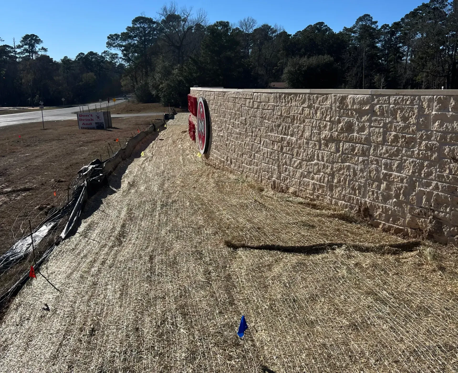 Curved beige retaining wall beside a dirt lot, with trees and open sky in the background.