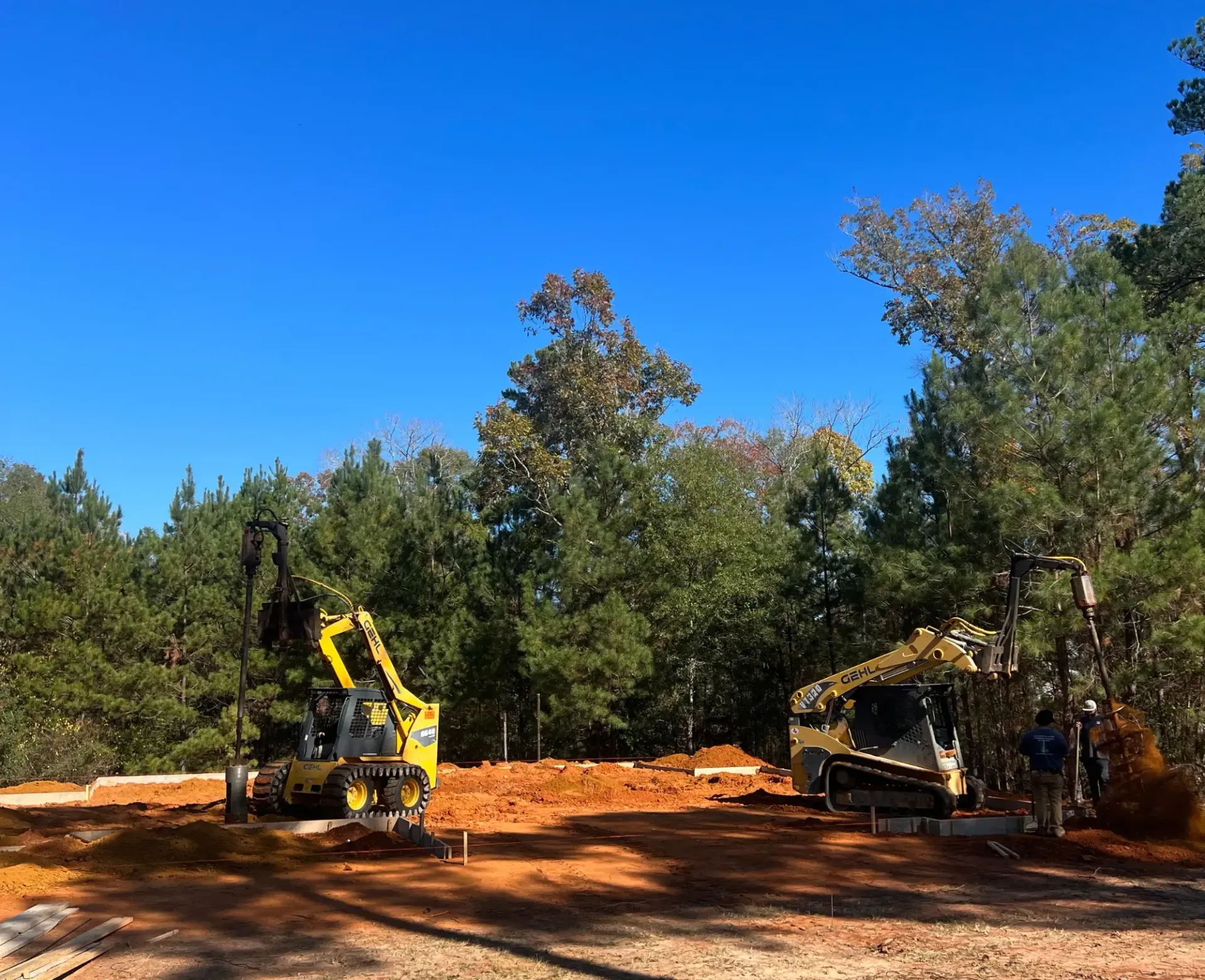 Construction site with excavators on a dirt clearing beneath a bright blue sky, surrounded by trees.