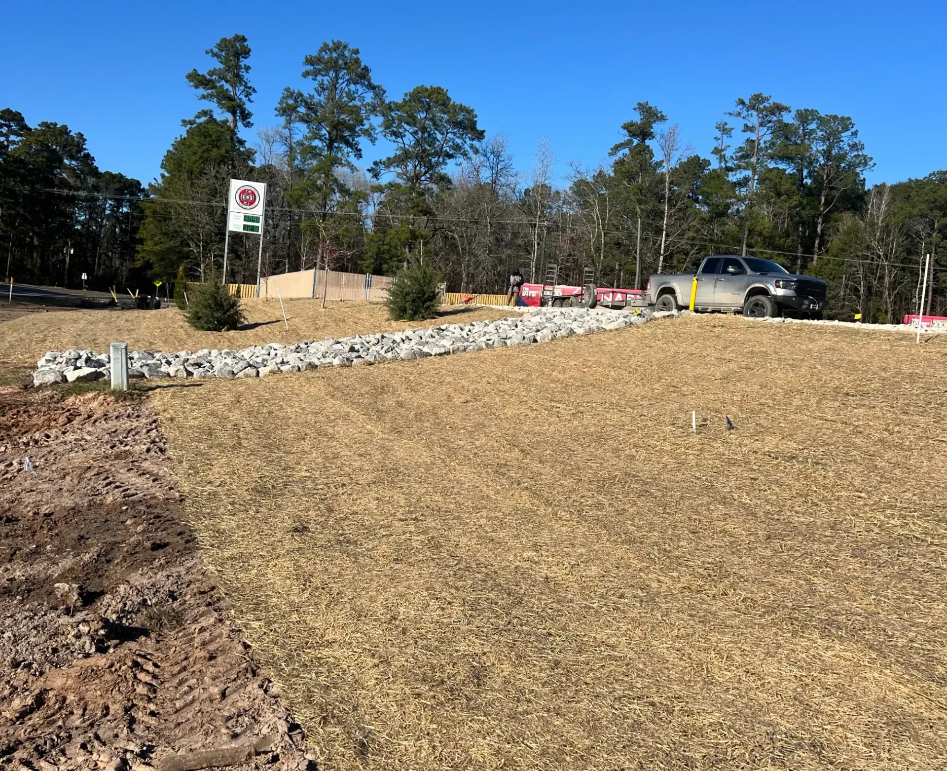 Gravel-covered lot under construction with parked vehicles, sign, and trees along the roadside