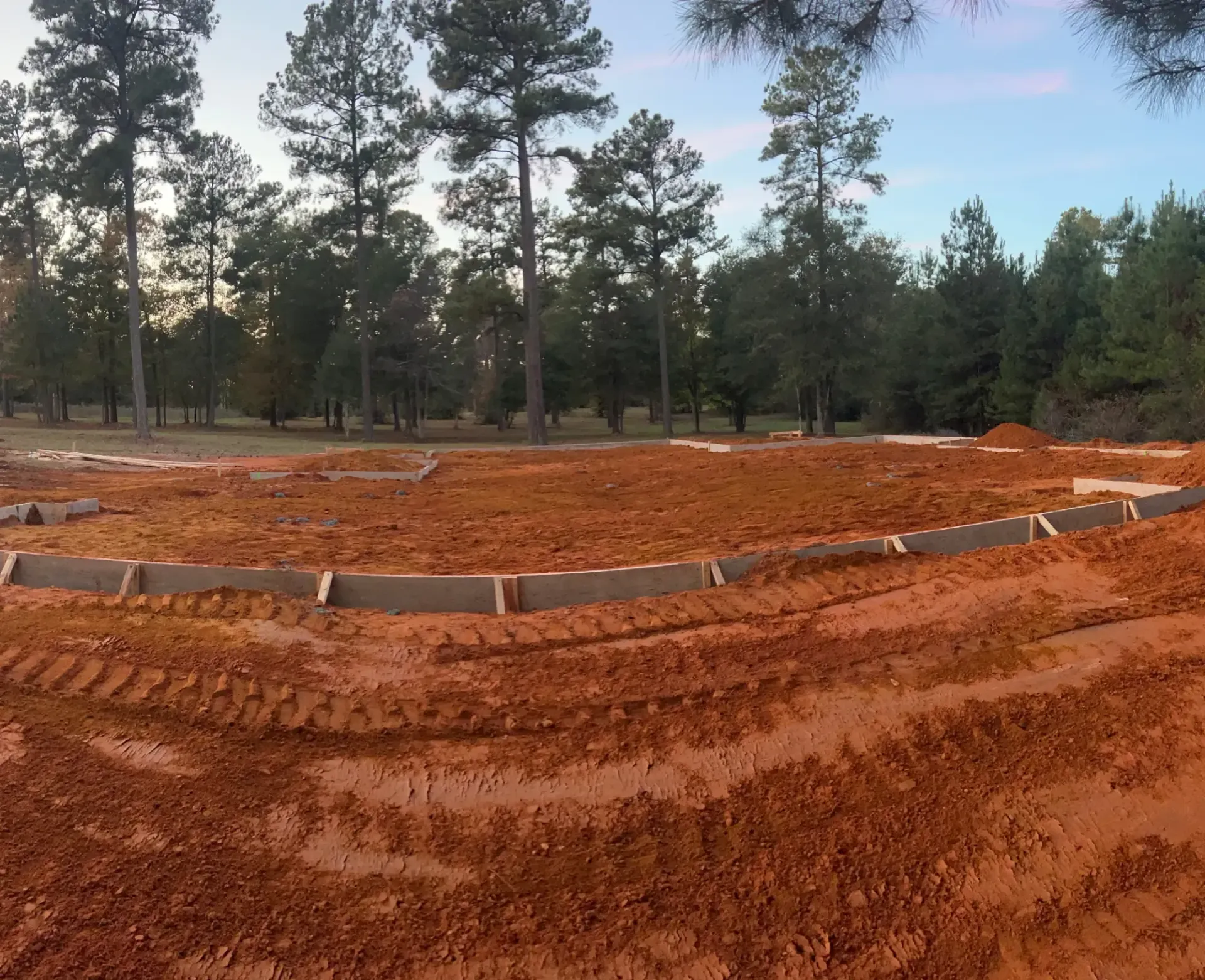Curved red dirt motocross track under construction, bordered by white blocks, with pine trees in the background