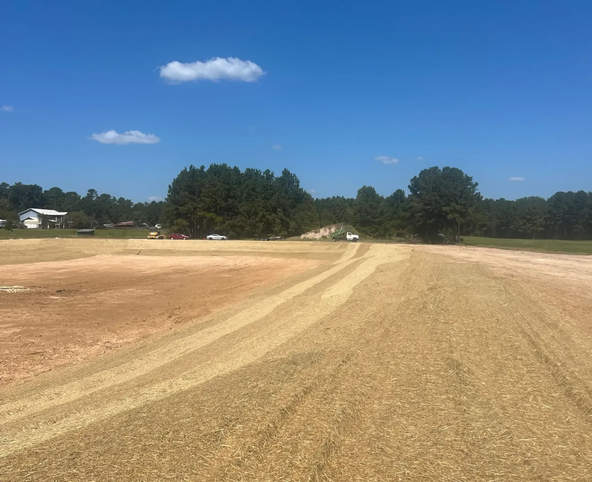 Freshly graded dirt field under a clear blue sky, with trees and houses in the distance.