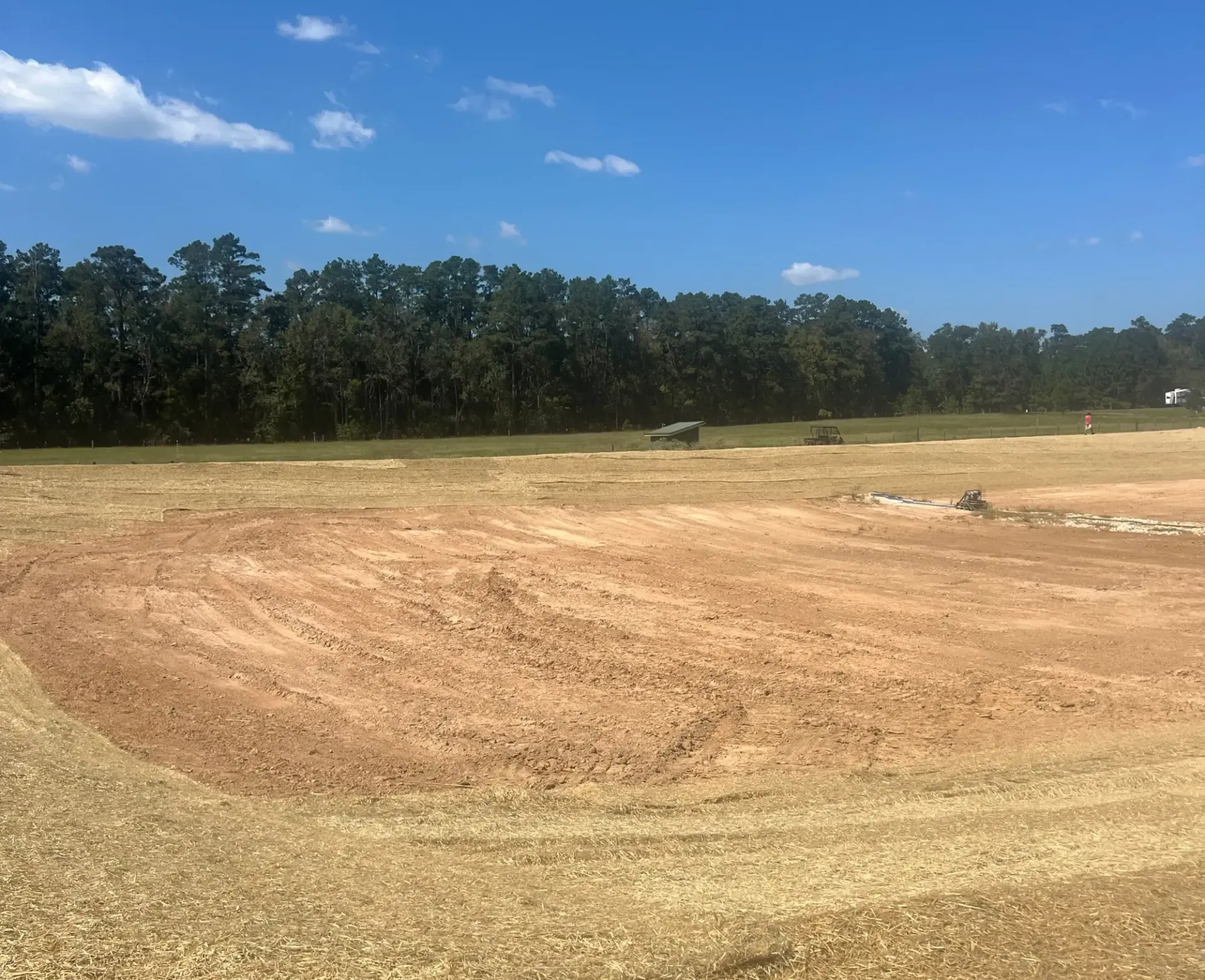 Open dirt field under blue sky, bordered by trees, with a small mound of debris in the distance