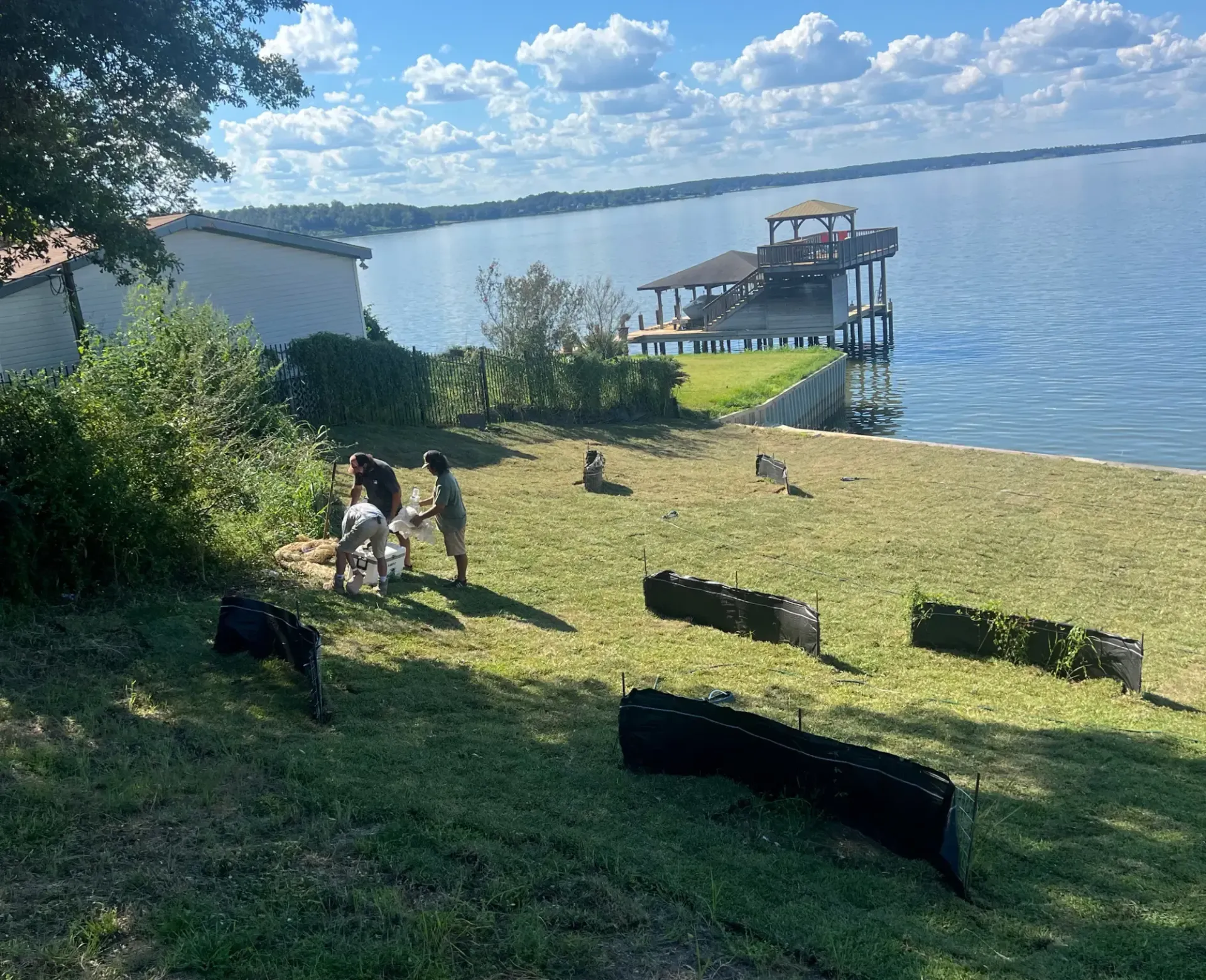 Sunny lakeside lawn with people sitting on the grass near a dock over calm water