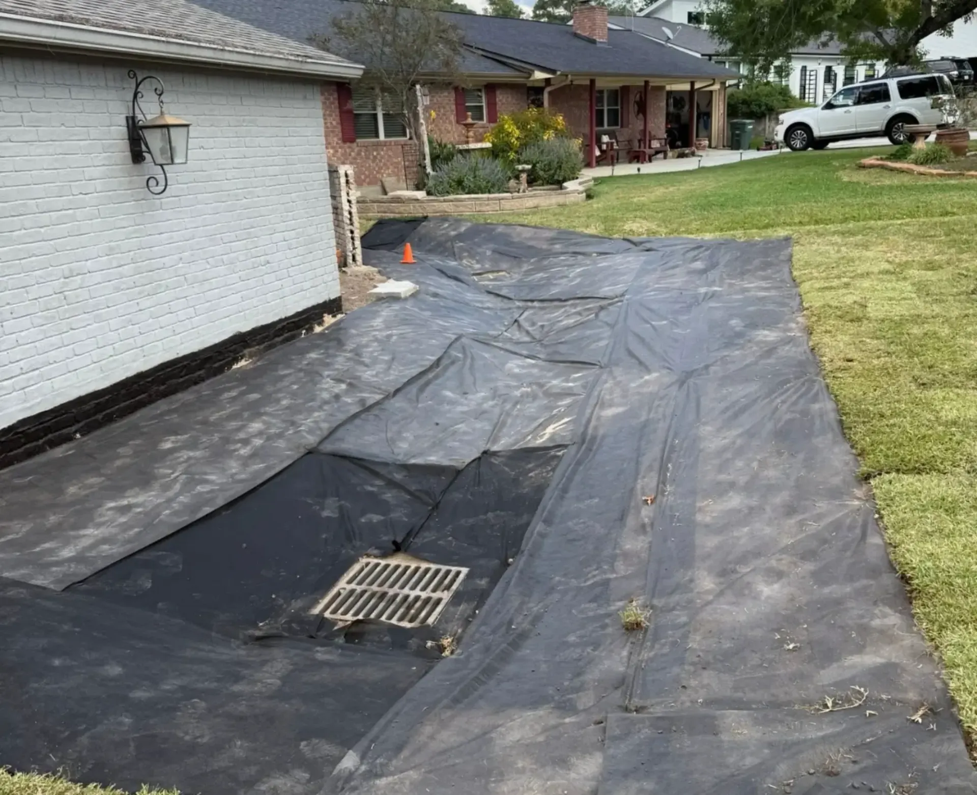 Black landscape fabric laid beside a white house, covering soil with a drain grate visible.
