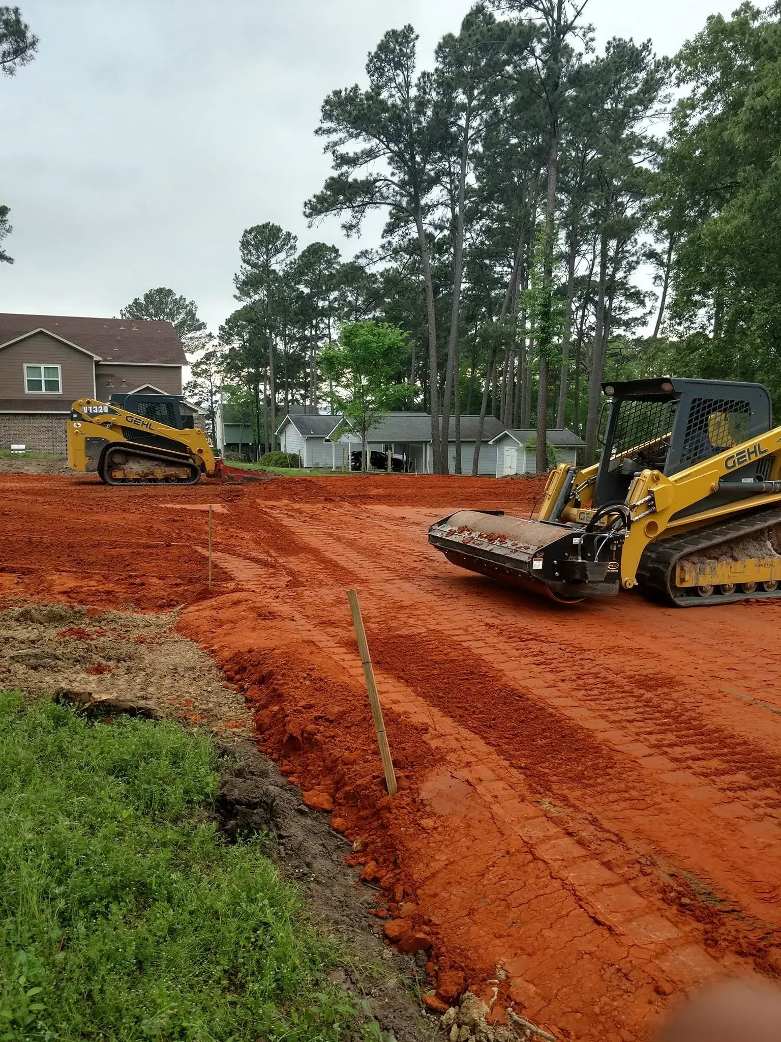 Yellow construction bulldozers grading a red dirt road on a residential lot with trees and houses nearby