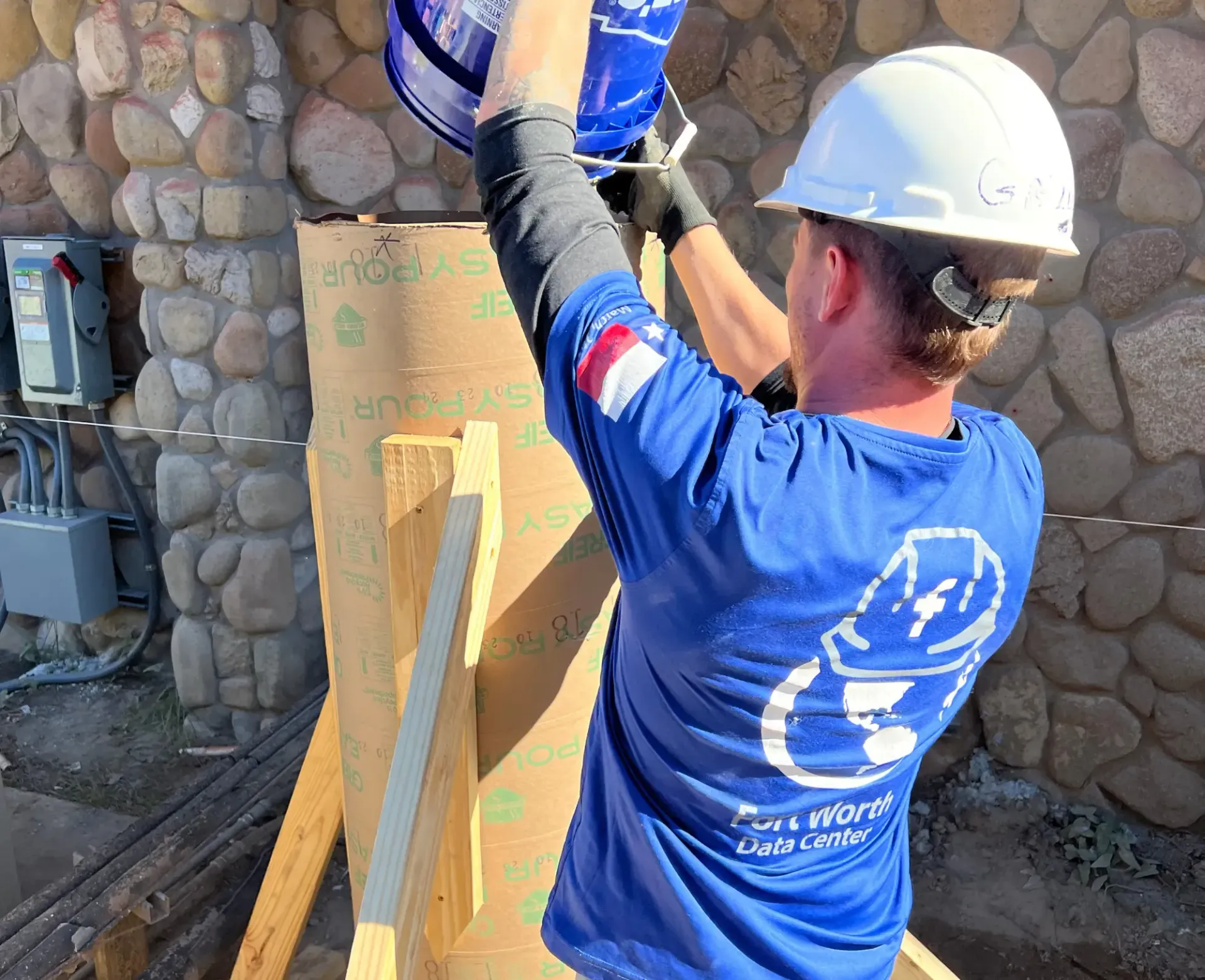 Worker in hard hat and blue shirt installing equipment against a stone wall outdoors.