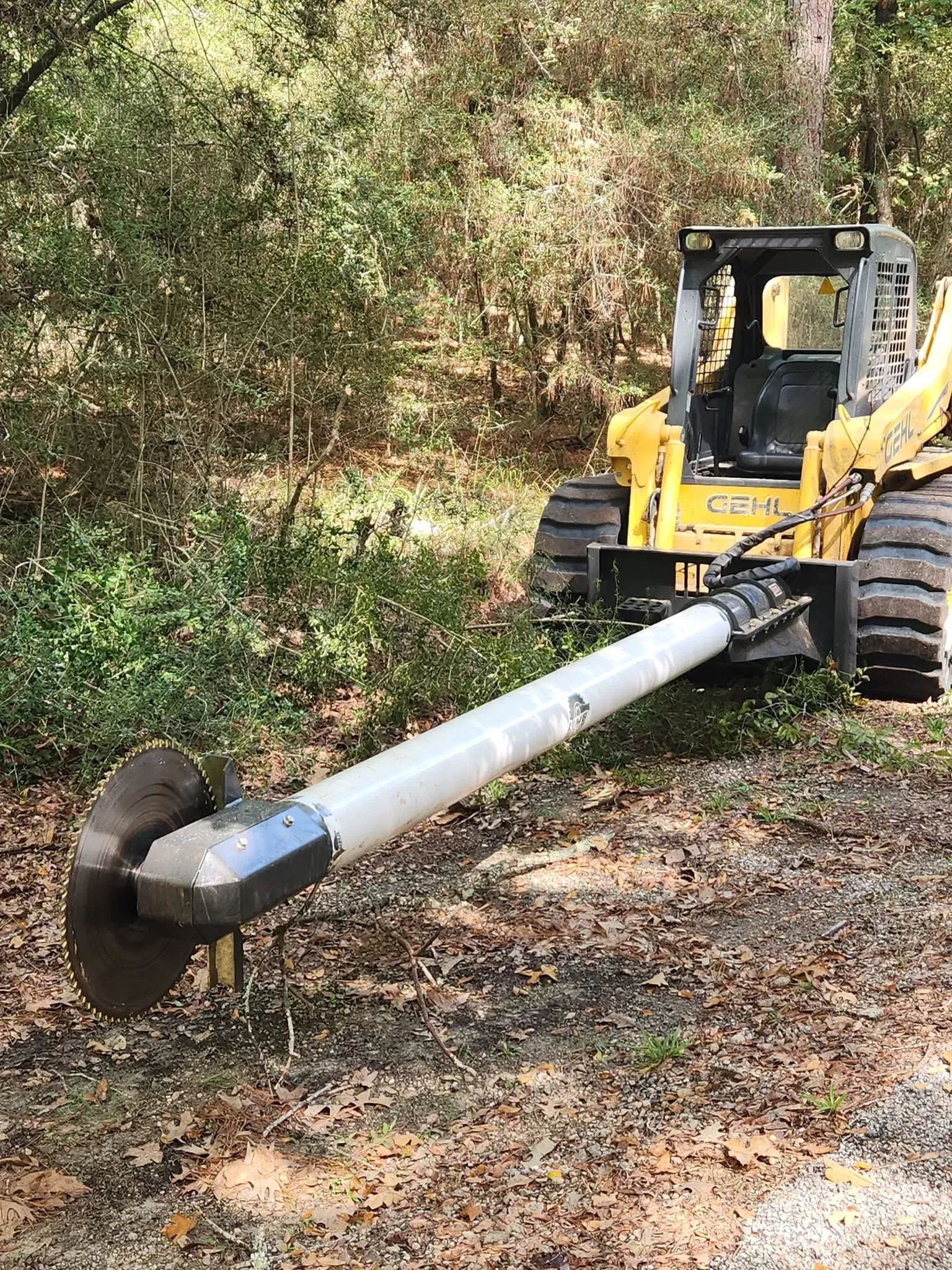 Yellow skid steer with a long auger attachment drilling into dirt beside a wooded roadside.