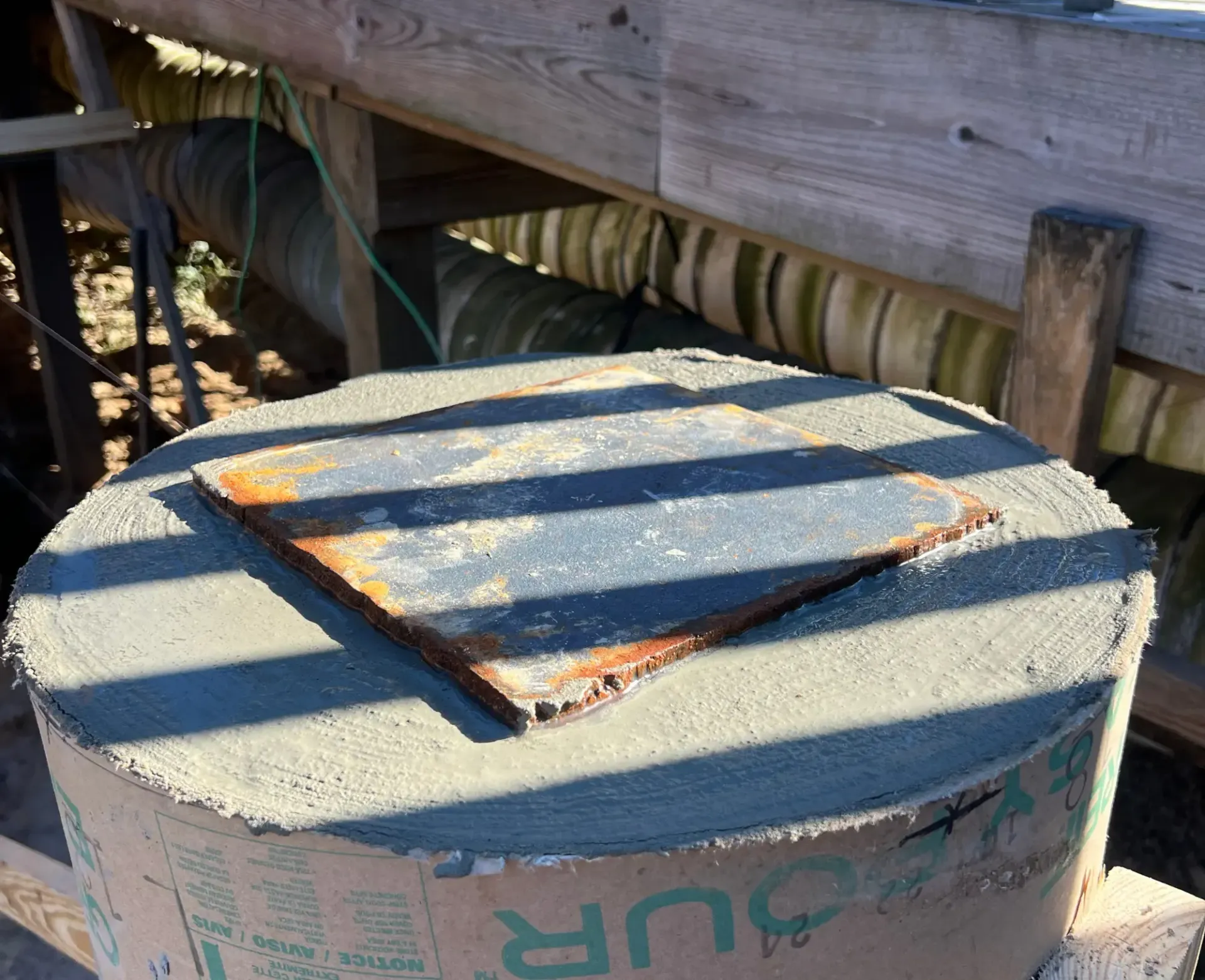 Round container lid with a rusty rectangular metal plate, outdoors in sunlight and shadow.