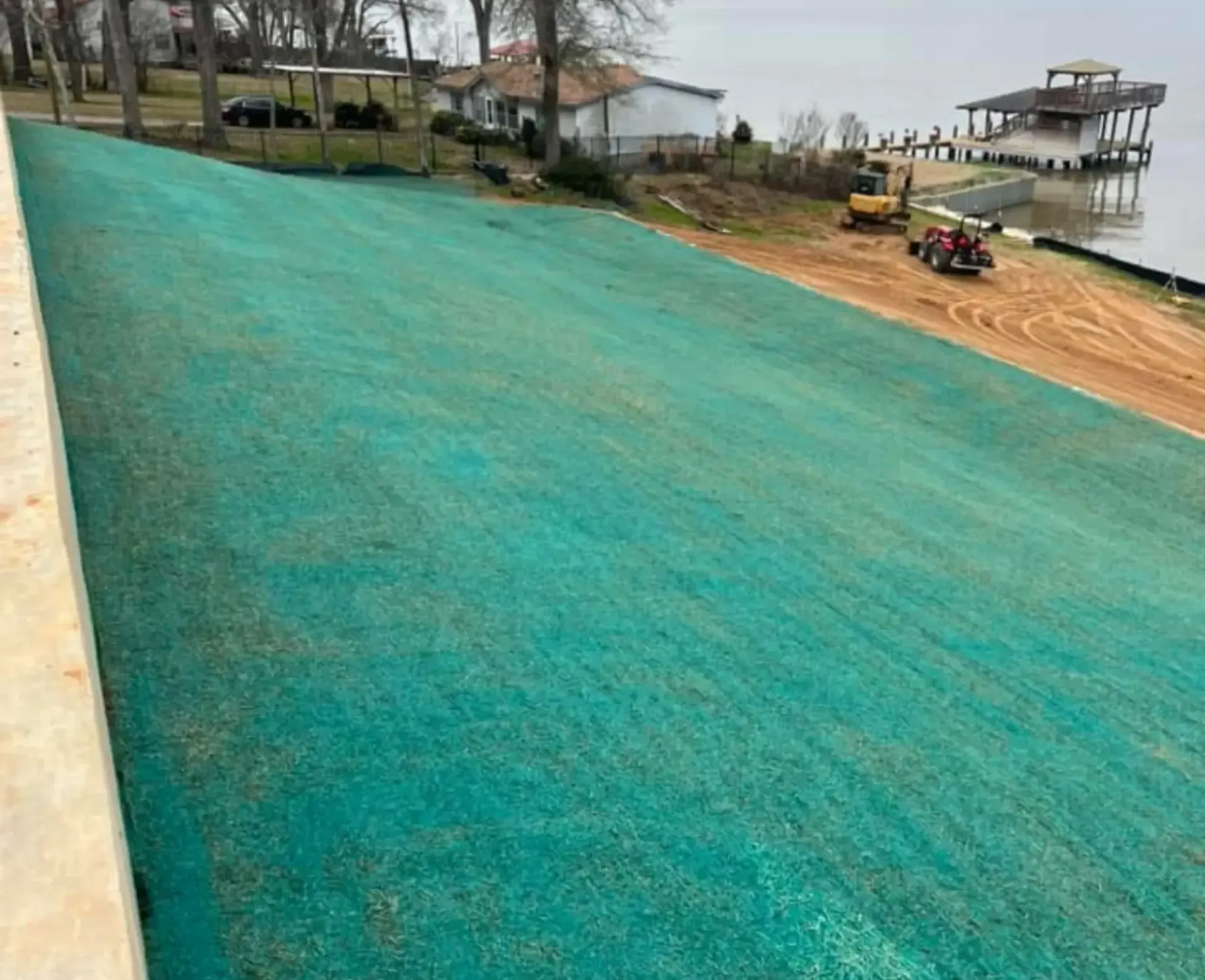 Freshly graded turquoise lawn beside a sandy lakeshore with construction equipment and a dock in the distance