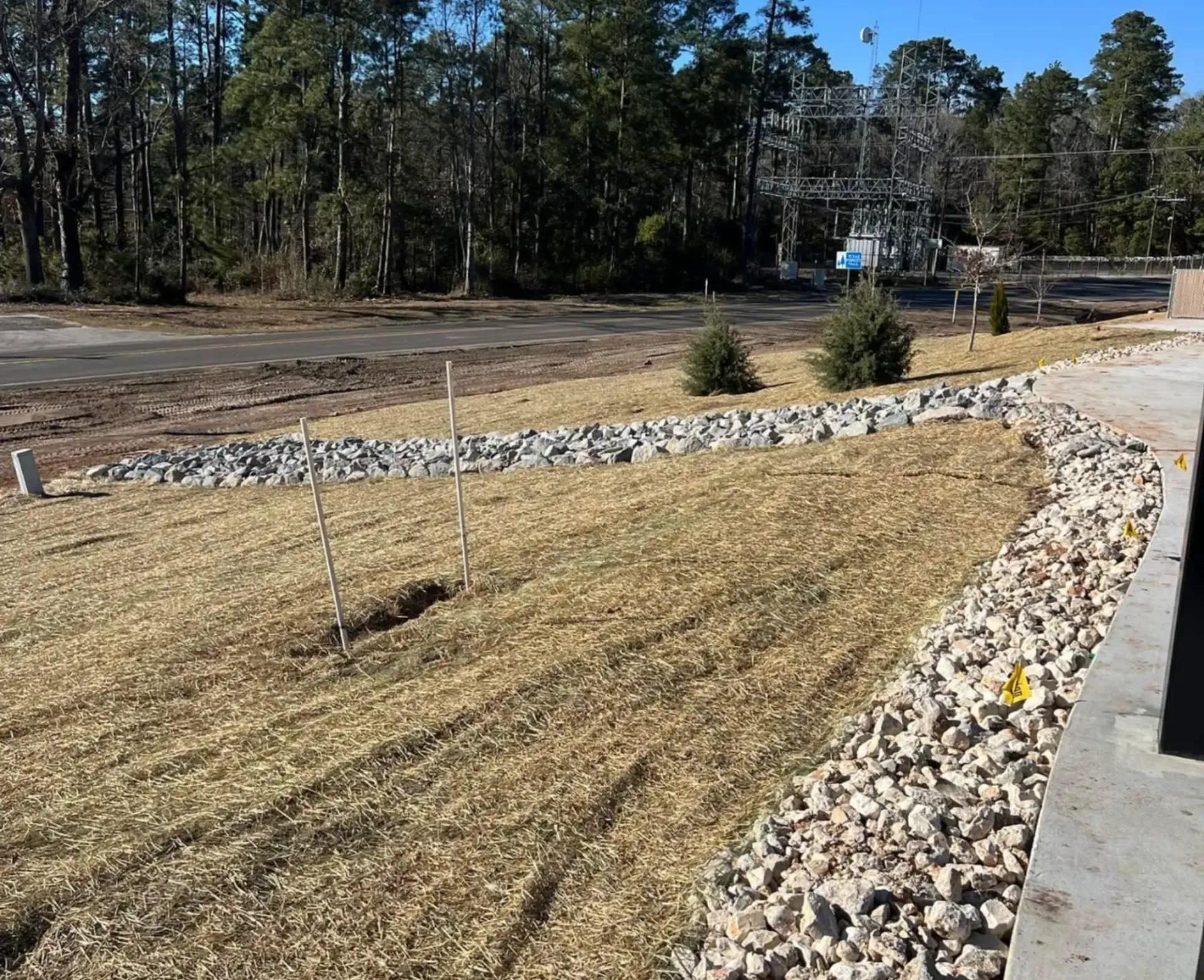 Mulched landscaped slope with rocks and young shrubs beside a paved path, trees and road in the background