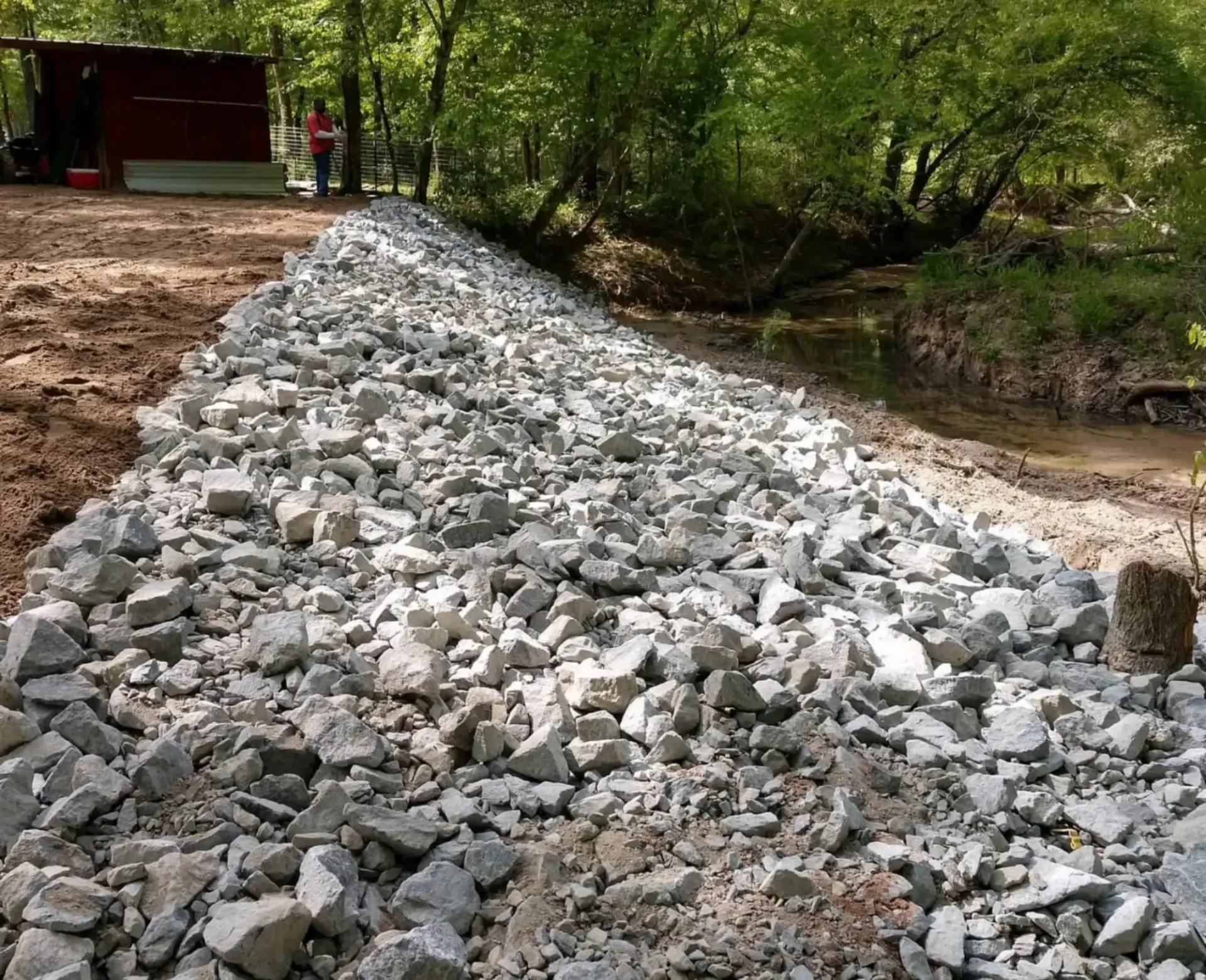 Rock-covered embankment beside a creek, with trees and a small building in the background.