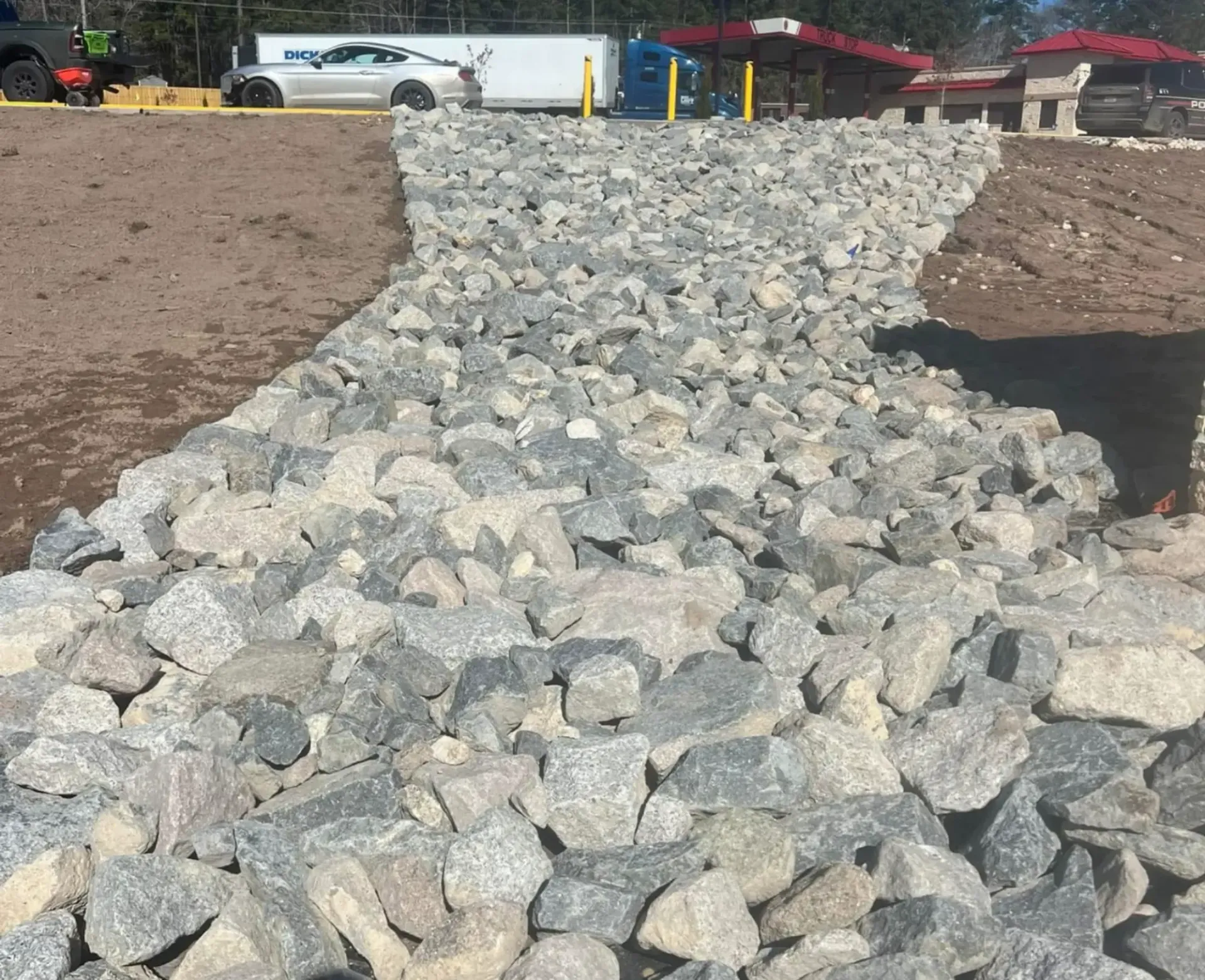 Rock-lined drainage channel cutting through a dirt construction site, with vehicles in the background