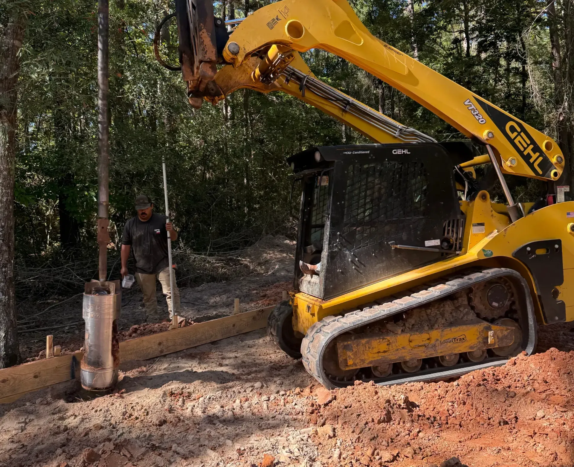 Yellow excavator clearing dirt beside a wooded path, with a worker nearby