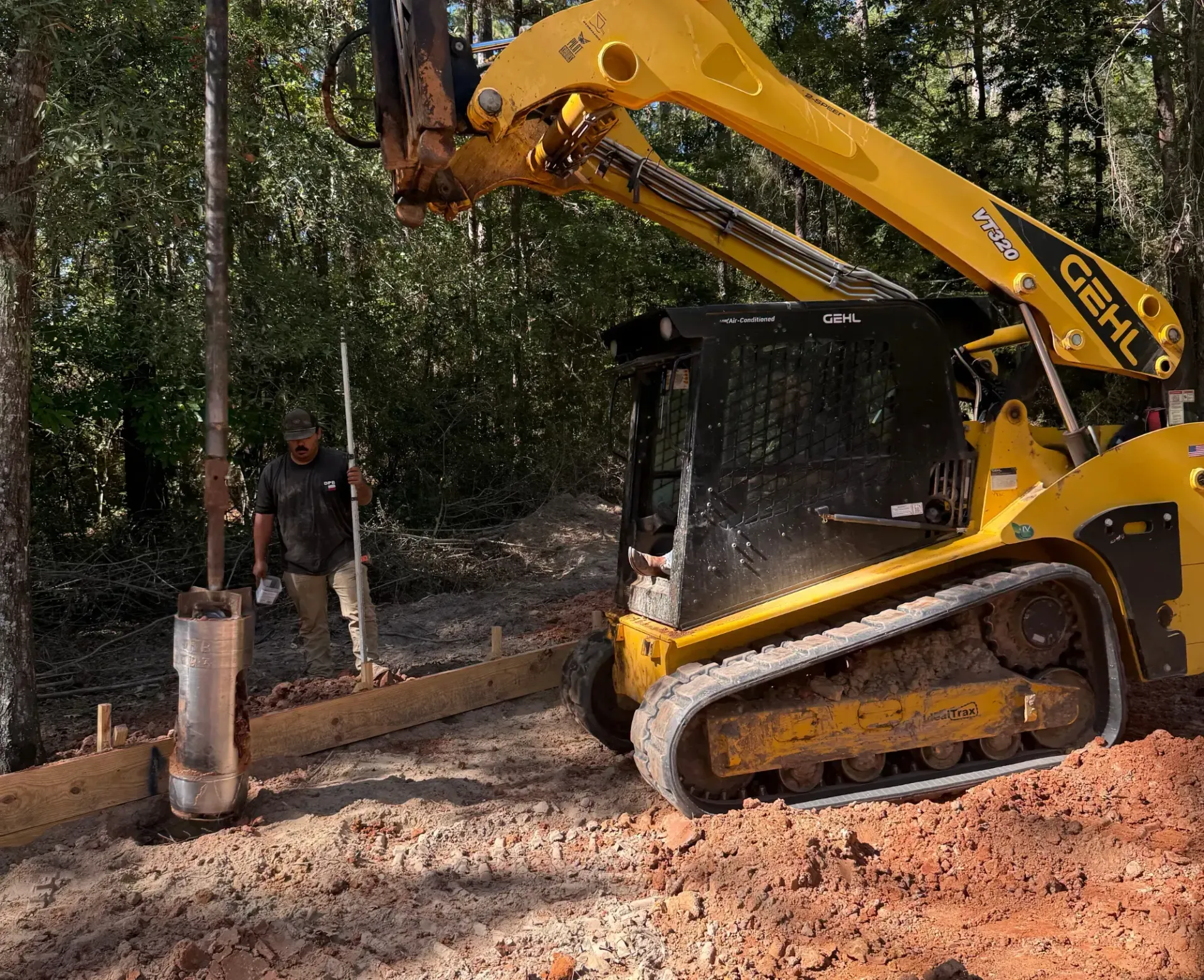 Yellow excavator at a wooded construction site, lifting a metal post while a worker stands nearby.