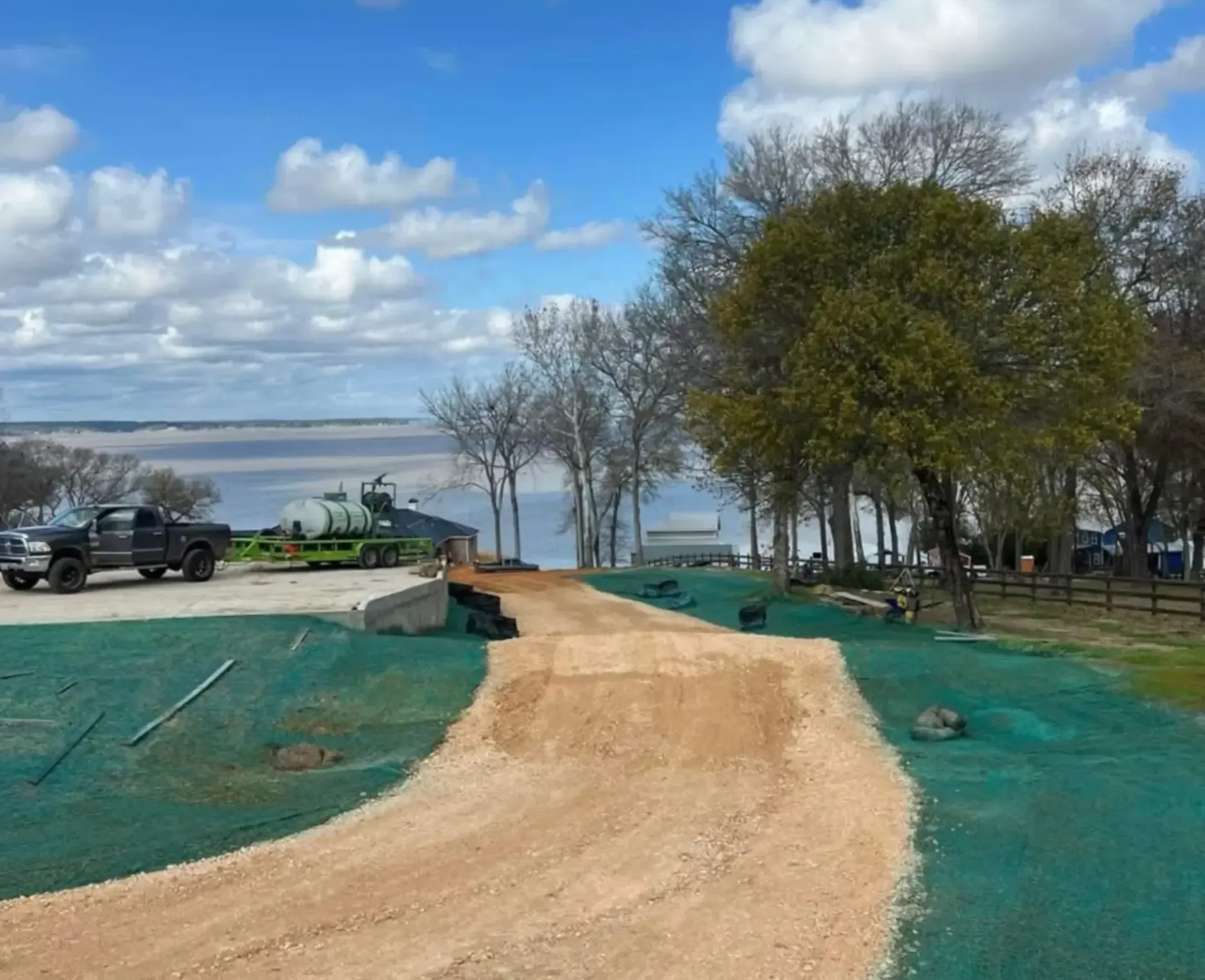 Dirt path leading to a lakeside area with trees, parked vehicles, and a partly cloudy sky