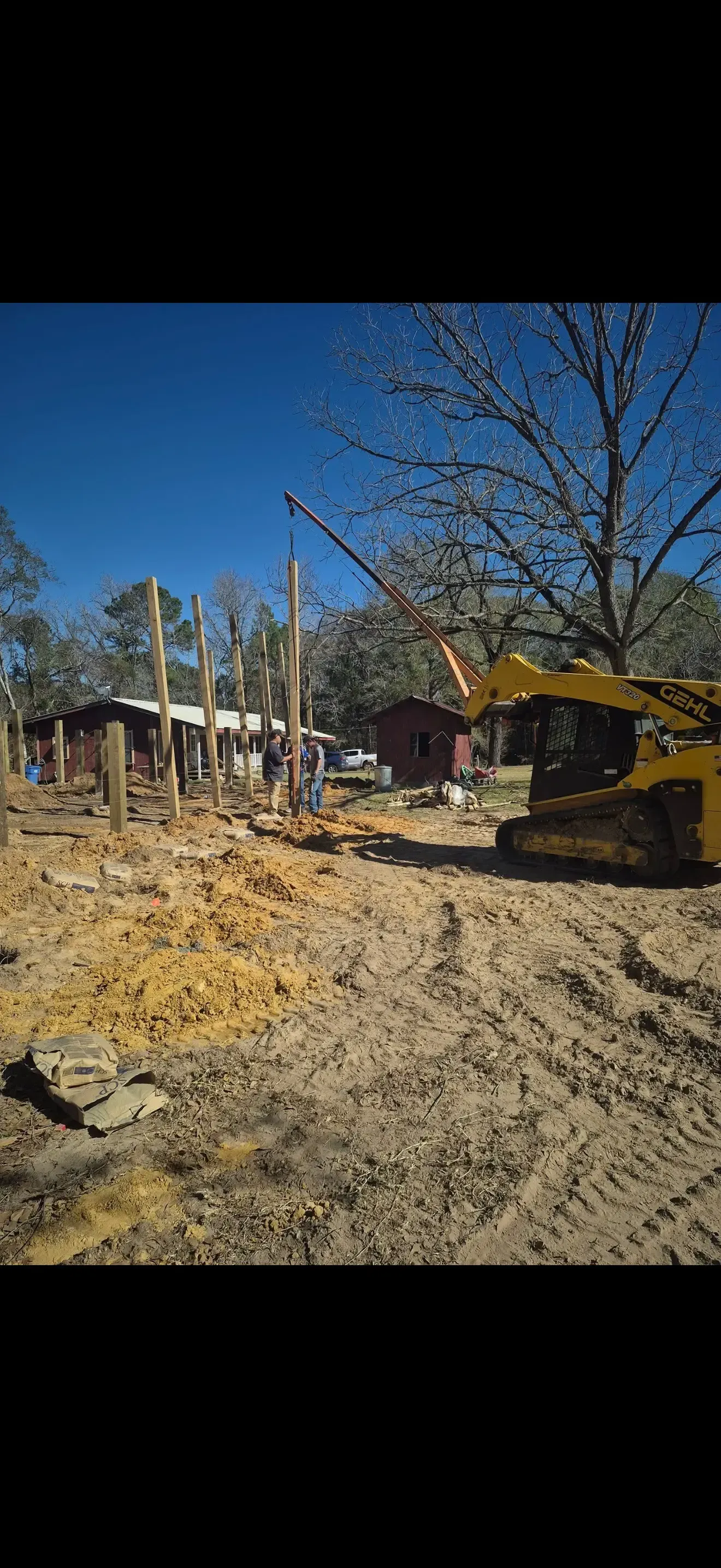 Excavator clearing dirt at a construction site under a blue sky, with trees and a house in the background
