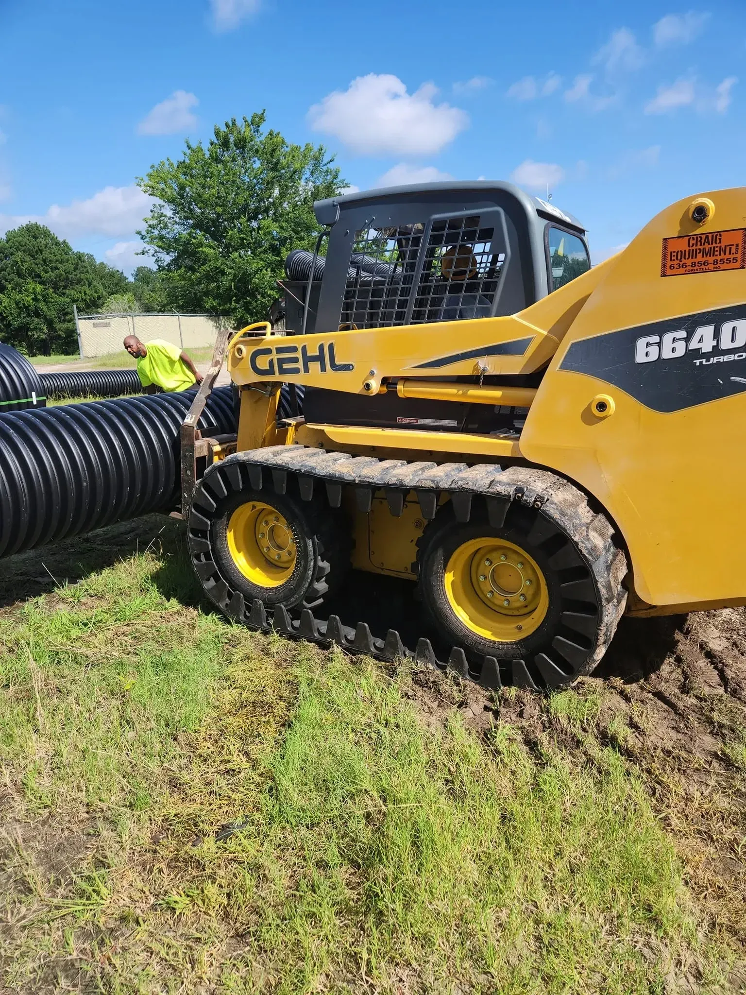 Yellow Gehl skid steer moving a large black pipe in a grassy field under a blue sky