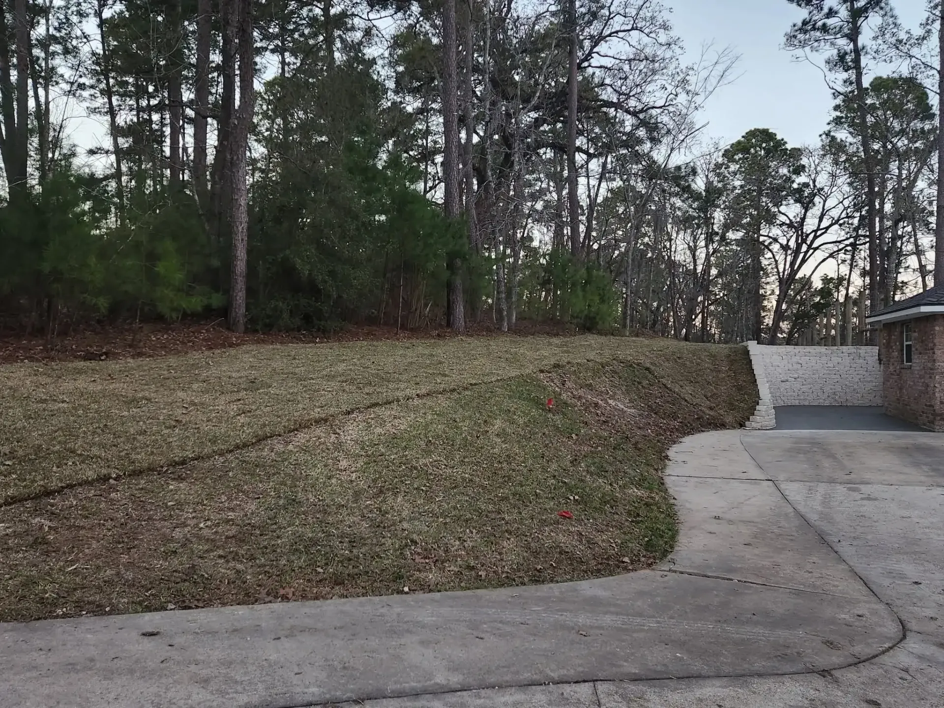 Backyard with stone retaining wall, brick building, and grassy slope near trees