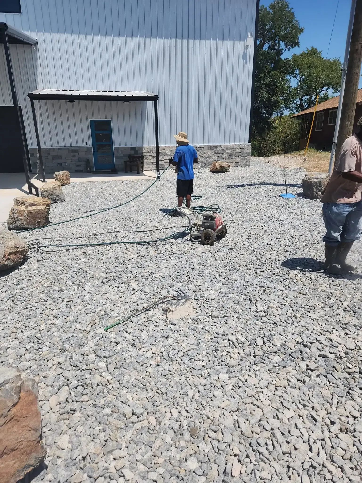 Two workers on a gravel lot beside a metal building, one in a blue shirt and hat, one holding a hose or cable.