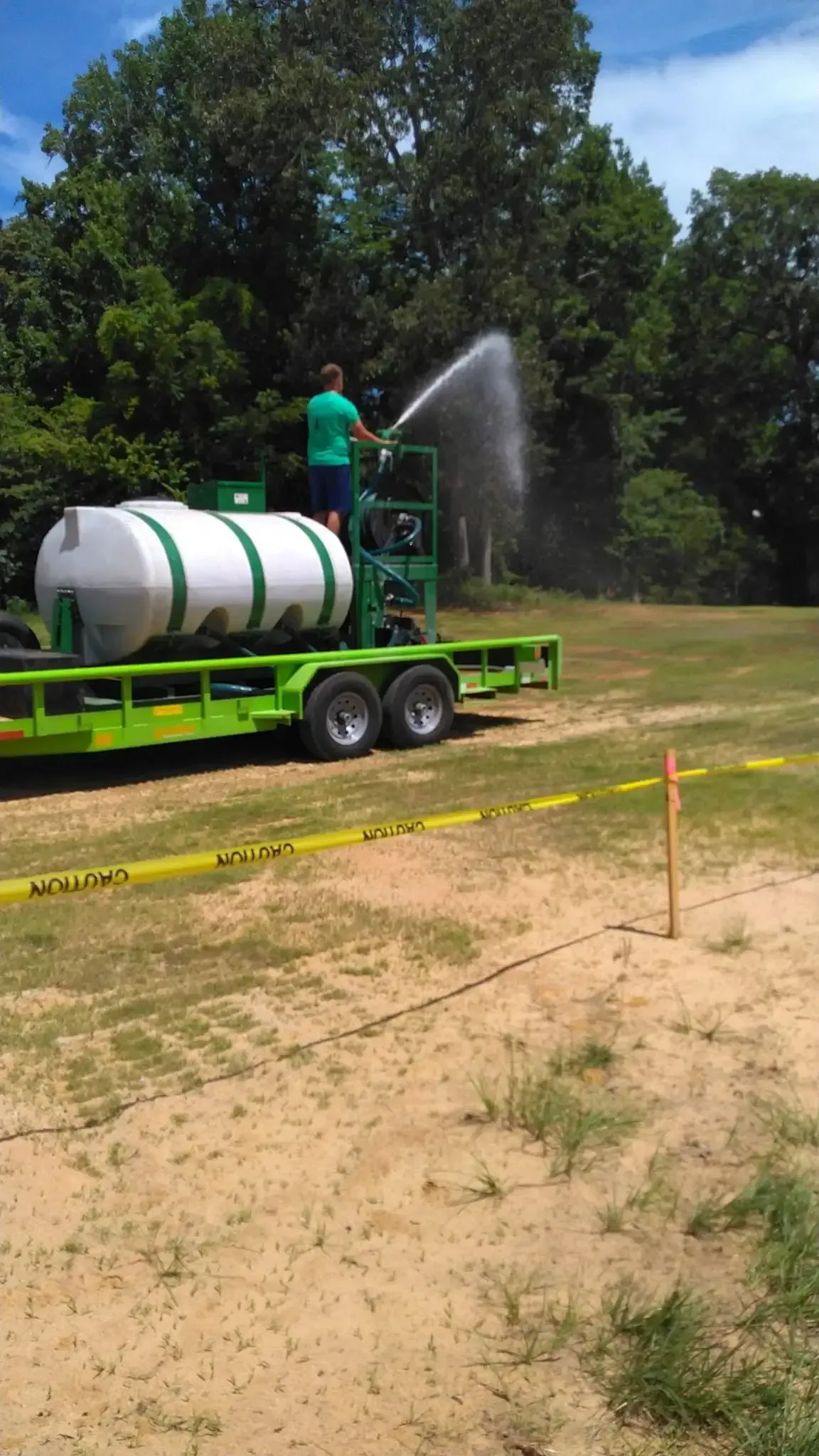 Green water tank trailer spraying water on a dirt field beside yellow caution tape under a blue sky