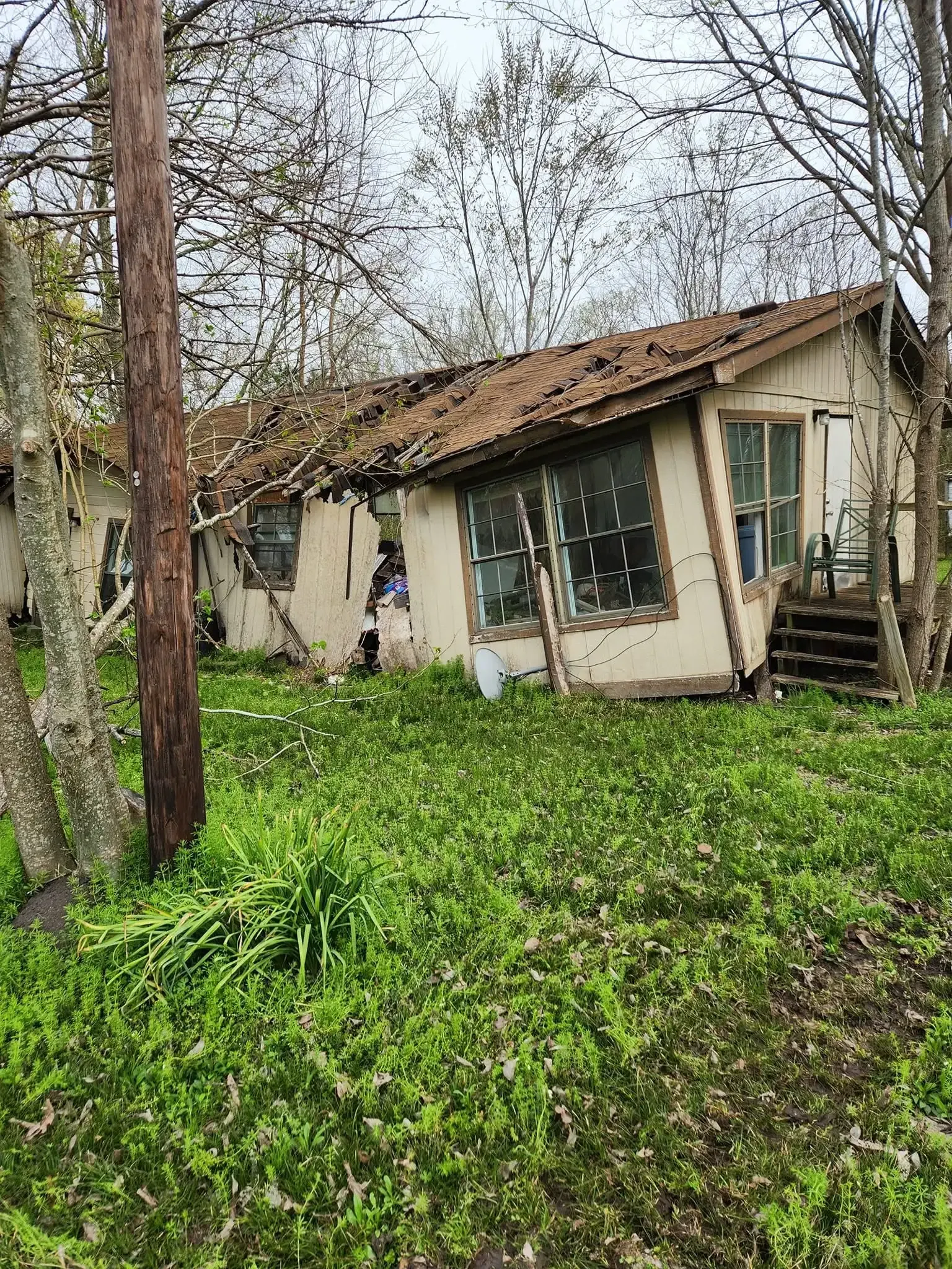 Dilapidated house with broken windows and a sagging roof, surrounded by overgrown grass and trees.