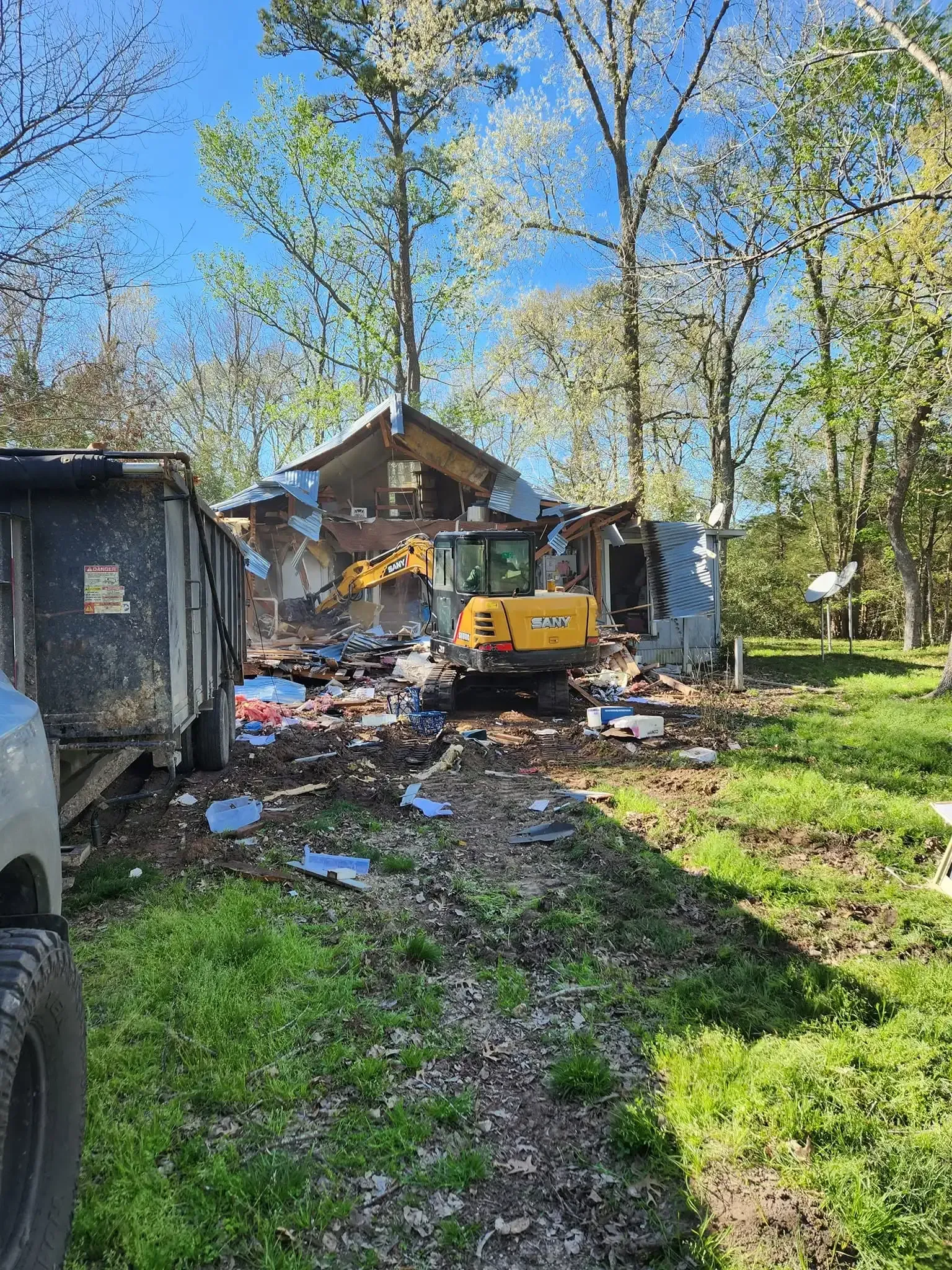Cluttered yard with debris, a yellow excavator, sheds, and tall trees under a clear blue sky