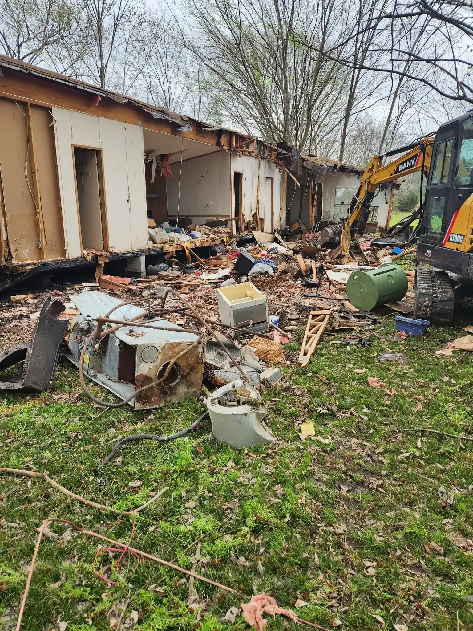 Collapsed house with debris scattered in a yard, and a damaged excavator beside the wreckage.