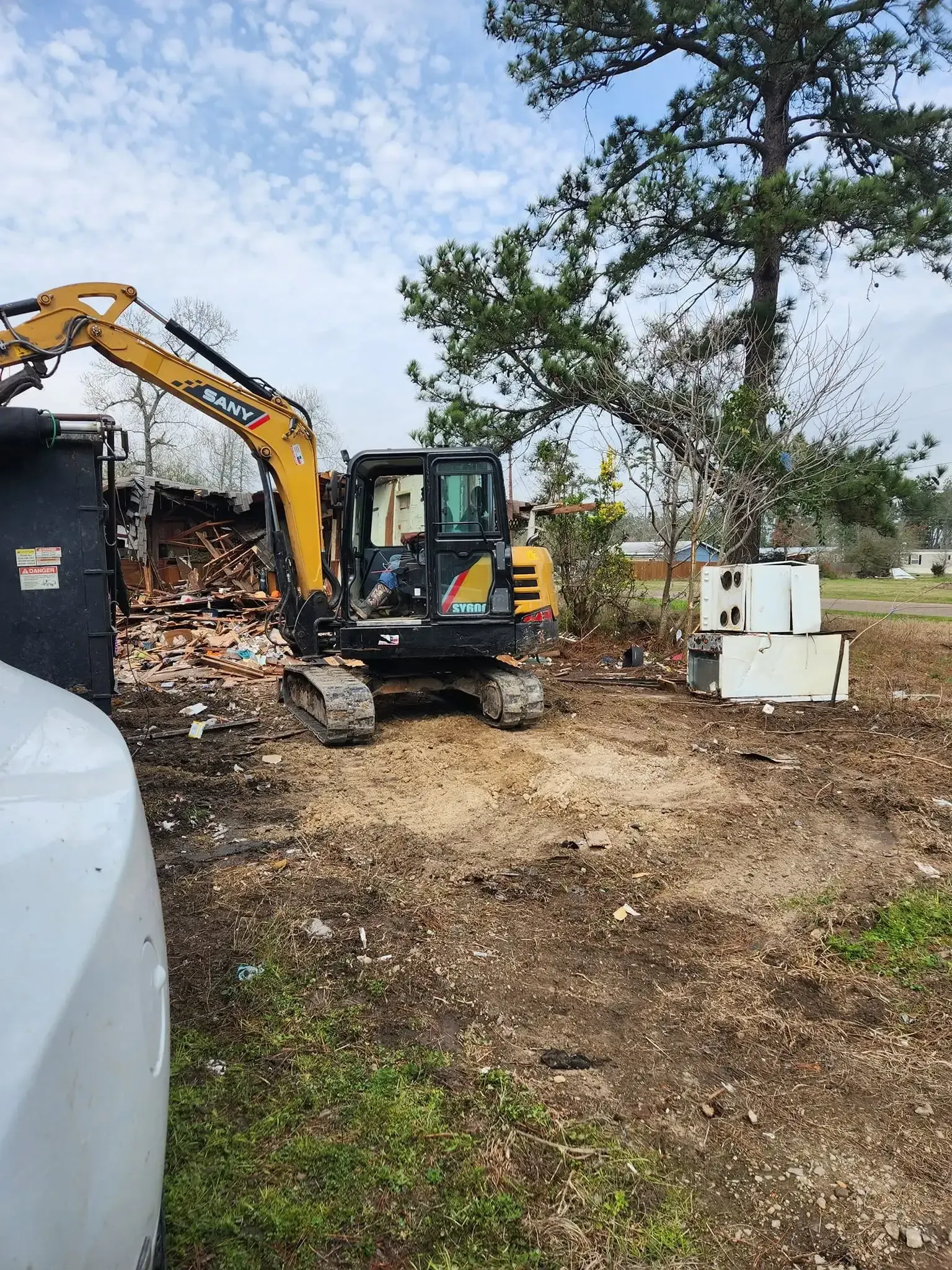 Excavator clearing debris at a construction site with trees and a cloudy sky behind it