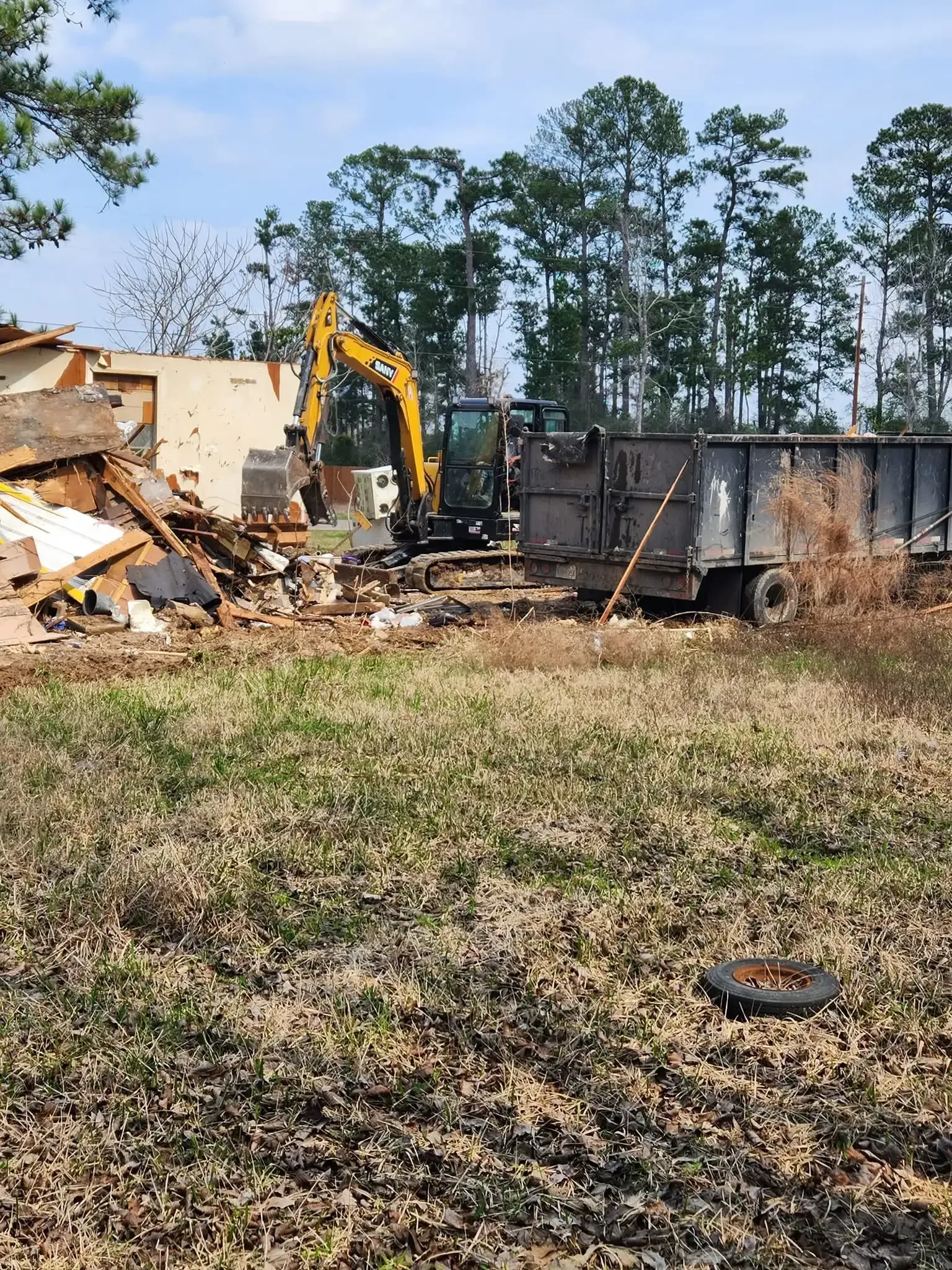 Yellow excavator demolishing debris beside a dumpster in a grassy lot under a clear sky