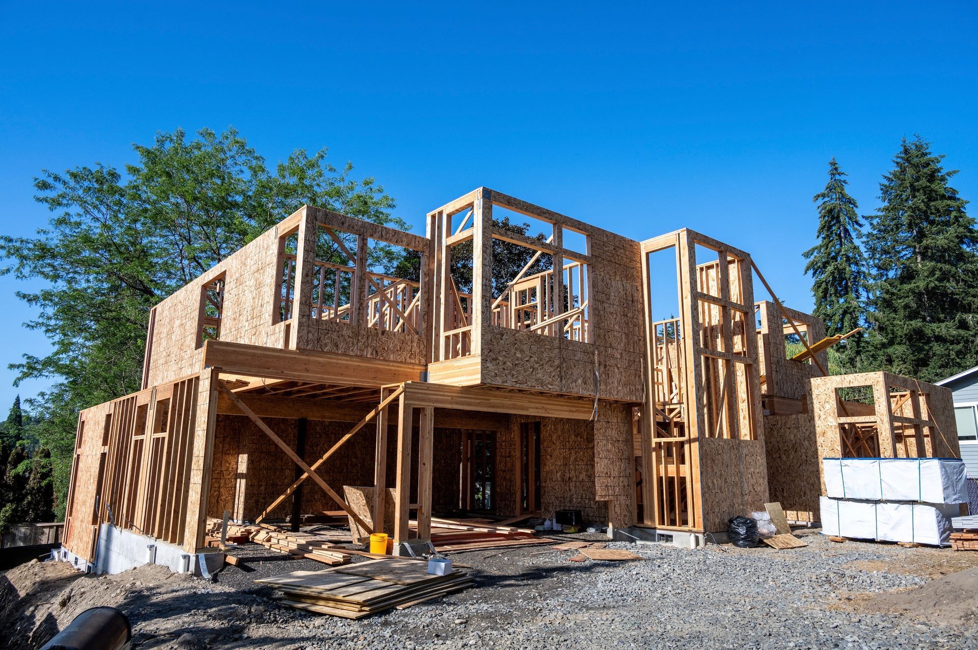 A two-story house under construction with exposed wooden framing and plywood sheathing on a sunny day.