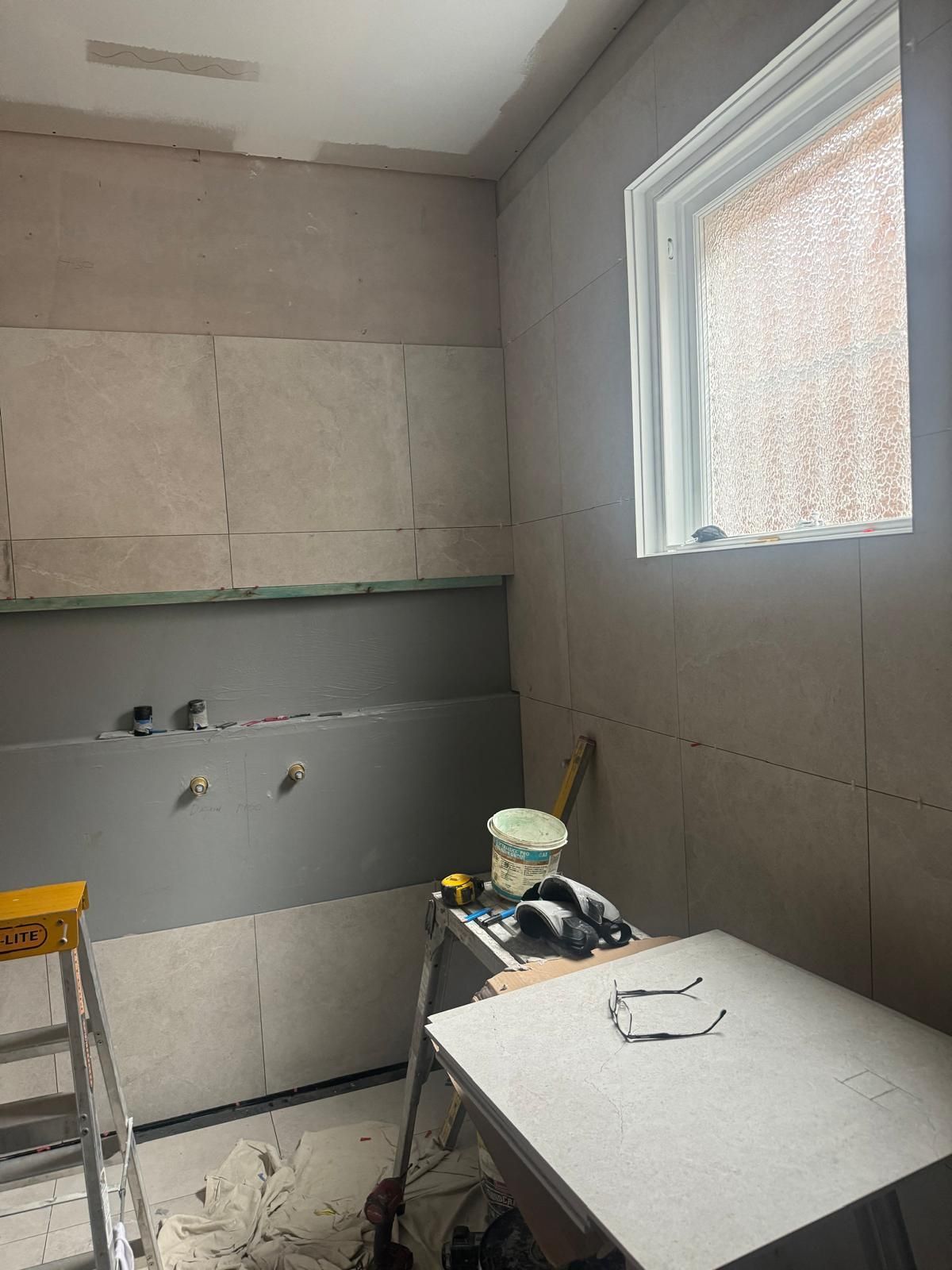 Bathroom under construction, drywall on walls, window, tools on a table, ladder, buckets.