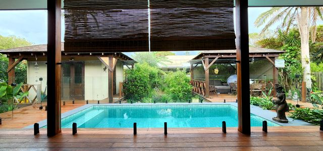 A view of a pool from a covered deck with a backyard and a building in the background.