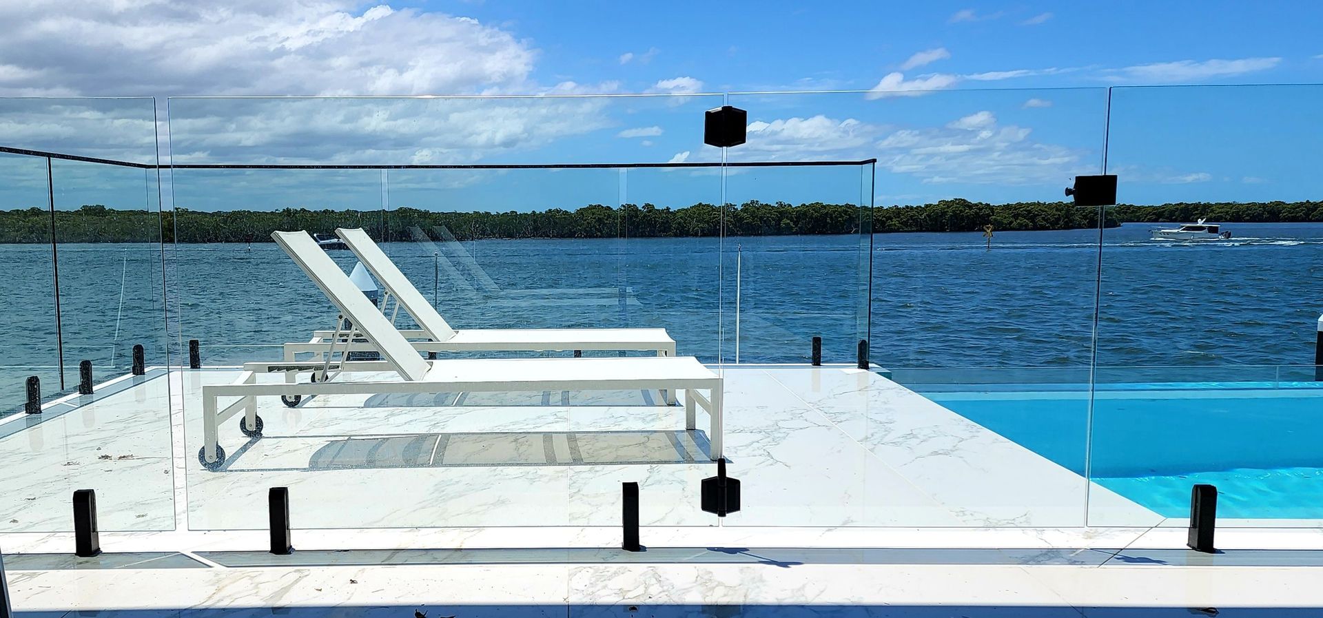 Two white lounge chairs on a white deck with glass railing overlooking the ocean, under a blue sky.